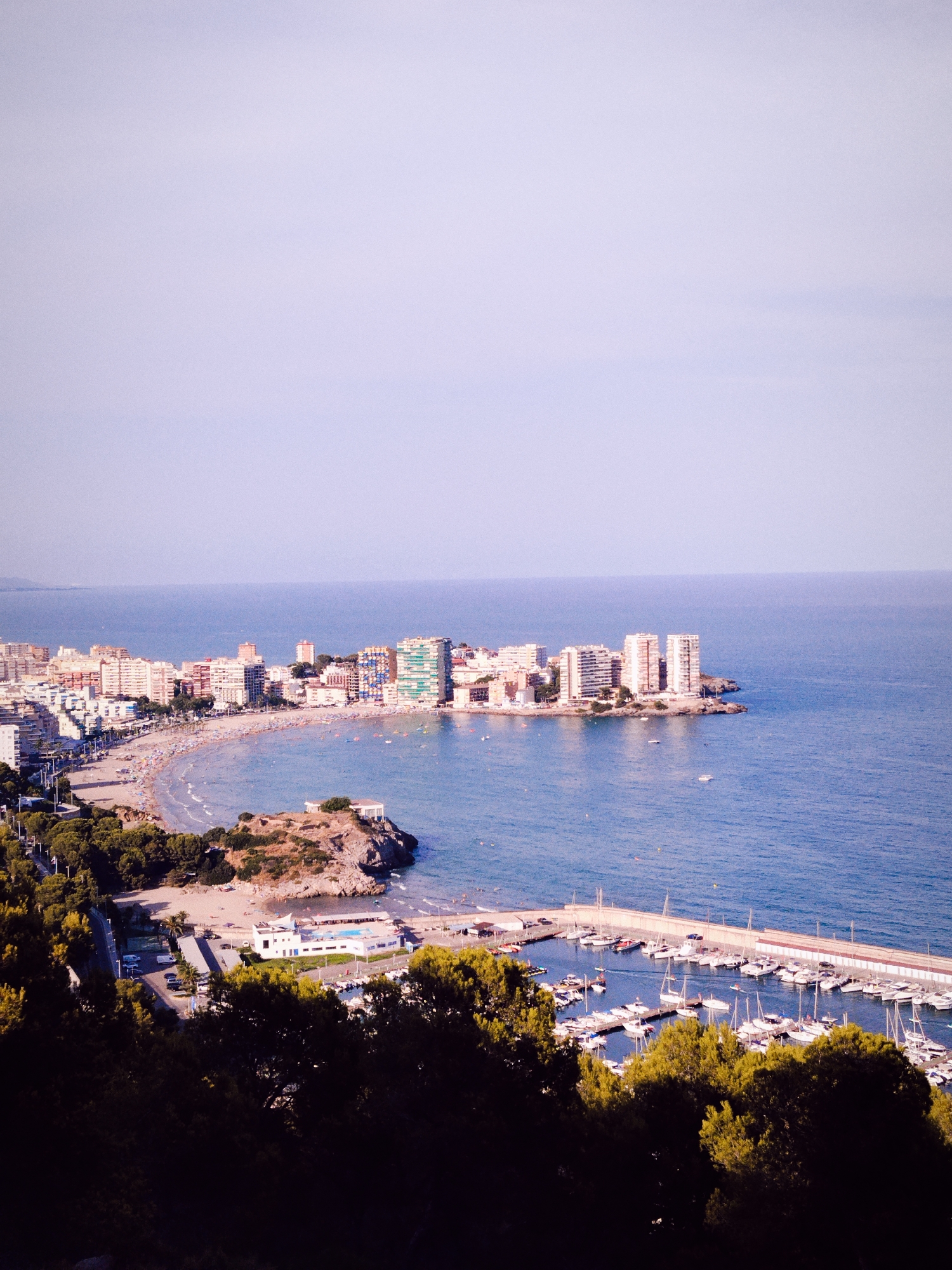 This image captures a panoramic view of the coastal city of Orpesa, Spain, showcasing its vibrant beachfront and bustling marina. The foreground is dominated by lush green trees, their dark silhouettes contrasting with the bright, sunlit scene beyond.
As the eye moves towards the midground, a crescent-shaped beach curves along the coastline, teeming with numerous beachgoers enjoying the sand and sea. Numerous buildings, ranging from apartment complexes to hotels, line the beach, their facades a mix of white, beige, and pastel colors. The architecture suggests a Mediterranean style, with many structures featuring balconies and large windows.
The turquoise waters of the Mediterranean Sea stretch out to the horizon, dotted with small boats and a larger marina filled with numerous sailboats and yachts. The marina is a prominent feature, with a long, white wall bordering a collection of moored vessels. The water is calm, reflecting the clear, pale blue sky.
On a rocky outcrop overlooking the beach, a white building with a flat roof and arched windows stands out, possibly a restaurant or a viewpoint. The overall scene conveys a sense of a popular tourist destination, alive with activity under a bright, sunny sky.