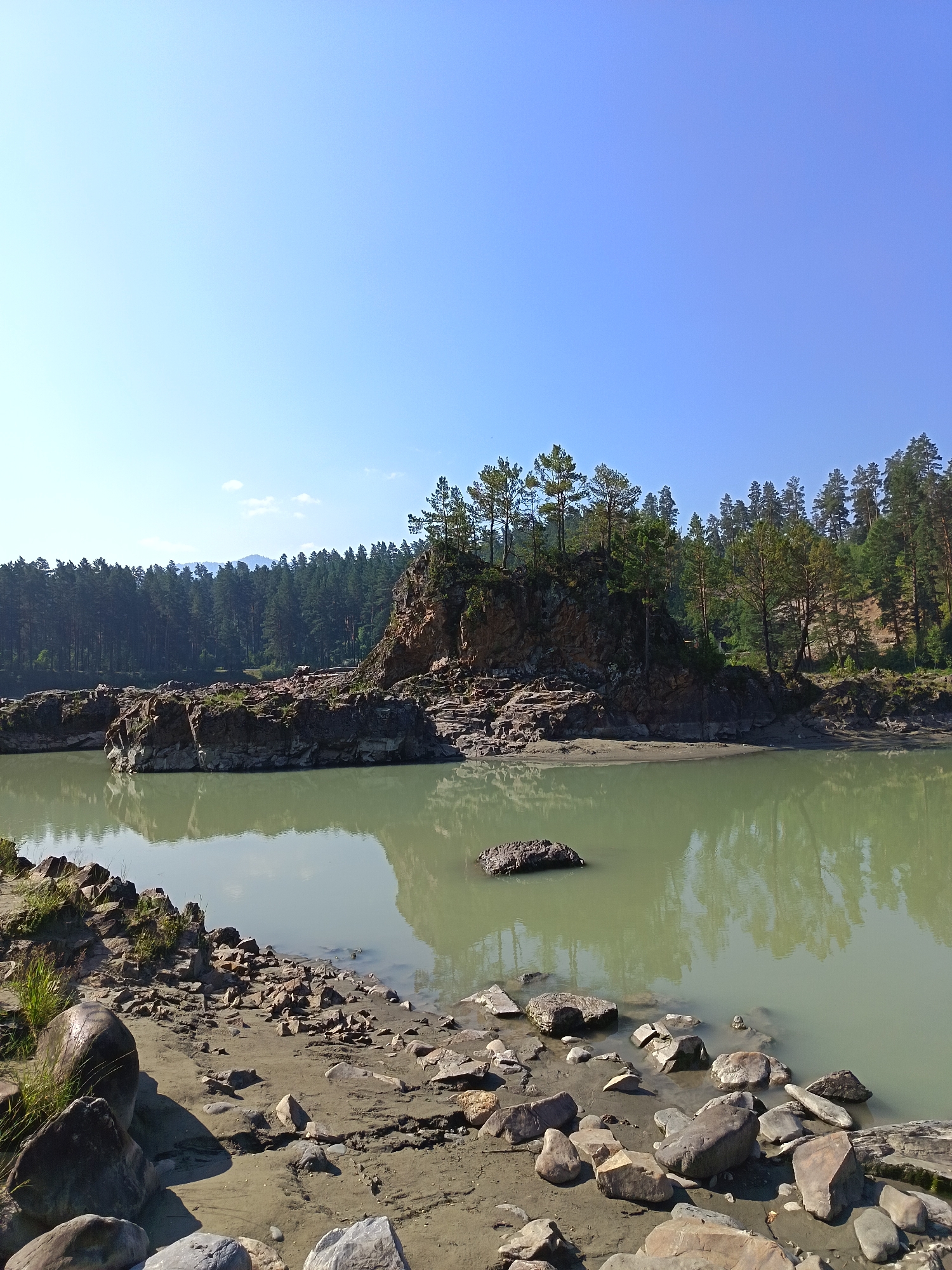 The image depicts a scenic natural landscape with a wide, tranquil river or lake in the foreground and a rocky, tree-covered island in the middle ground. The water is a pale green color and reflects the surrounding scenery. The island features rugged rock formations and is topped with a cluster of evergreen trees. Behind the island, a dense forest of pine trees covers a hillside. The sky above is a clear, bright blue. The riverbank in the foreground is composed of sand and various sized rocks. The overall impression is one of a serene and untouched natural environment. This image is likely taken in or near Manzherok, Russia, known for its natural beauty.