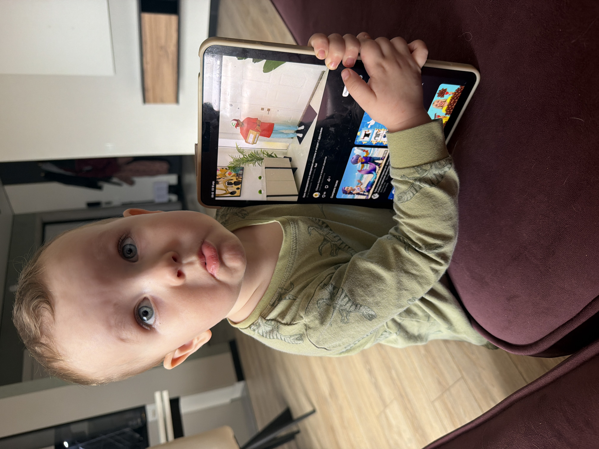 A young child with light hair and blue eyes holds a tablet in their hands. The tablet is displaying a video with a cartoon man wearing a red shirt and delivering a package. The video appears to be a cartoon or animation tutorial for home décor or renovation. The child is wearing a green long-sleeved shirt with dinosaur designs and appears to be sitting on a burgundy-colored chair. The background includes a modern interior design with wooden floors and neutral-colored furniture. The photo does not have sufficient information to determine the exact location in the city of Chaikovsky, Russia.