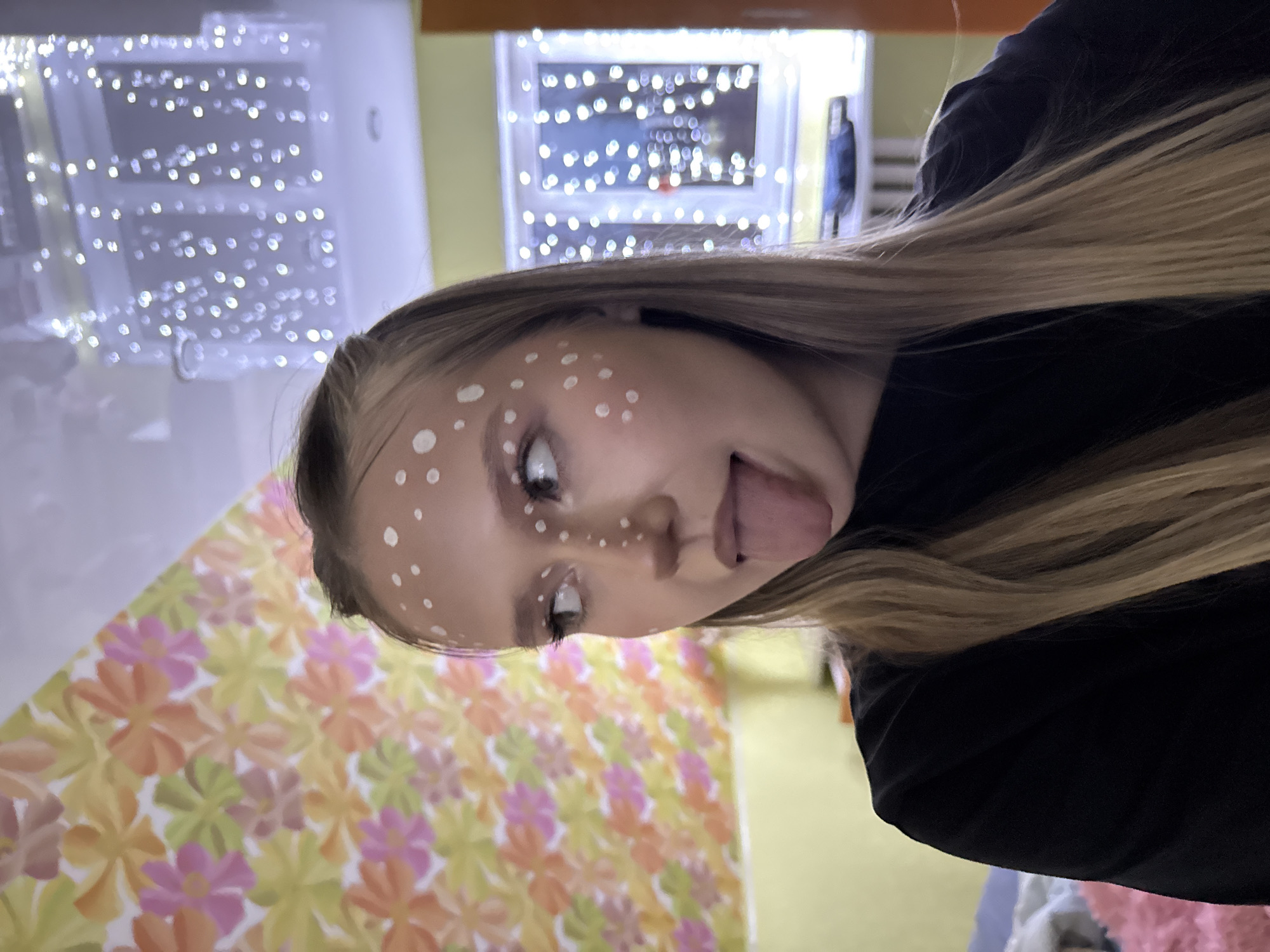 A young woman with blonde hair, facing slightly upward with her tongue sticking out, is the primary subject. White dots are painted on her forehead, nose, and cheeks. She is wearing a black garment.
Behind her, a wall is covered in a floral pattern of yellow, orange, and pink. Above her is a window with a string of white lights. Another wall is visible with similar strings of lights.
The location is Zhigalki, Russia.