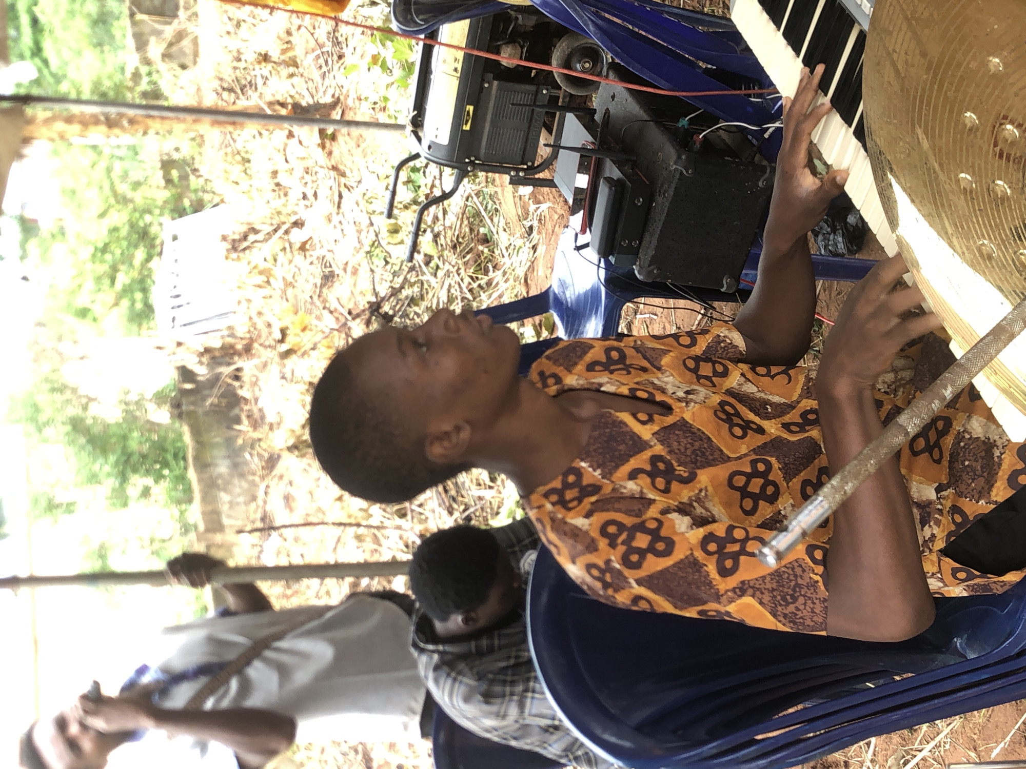 The image depicts a scene in Ukwu-Abwa, Nigeria. A dark-skinned person wearing an orange patterned shirt is seated and playing a keyboard. Another individual is positioned behind them, and a third is partially visible to the left. A cymbal and percussion instrument are in the foreground. The musical equipment includes speakers and a power generator situated on a blue plastic chair. The setting appears to be outdoors, as foliage and dry grass are seen in the background.