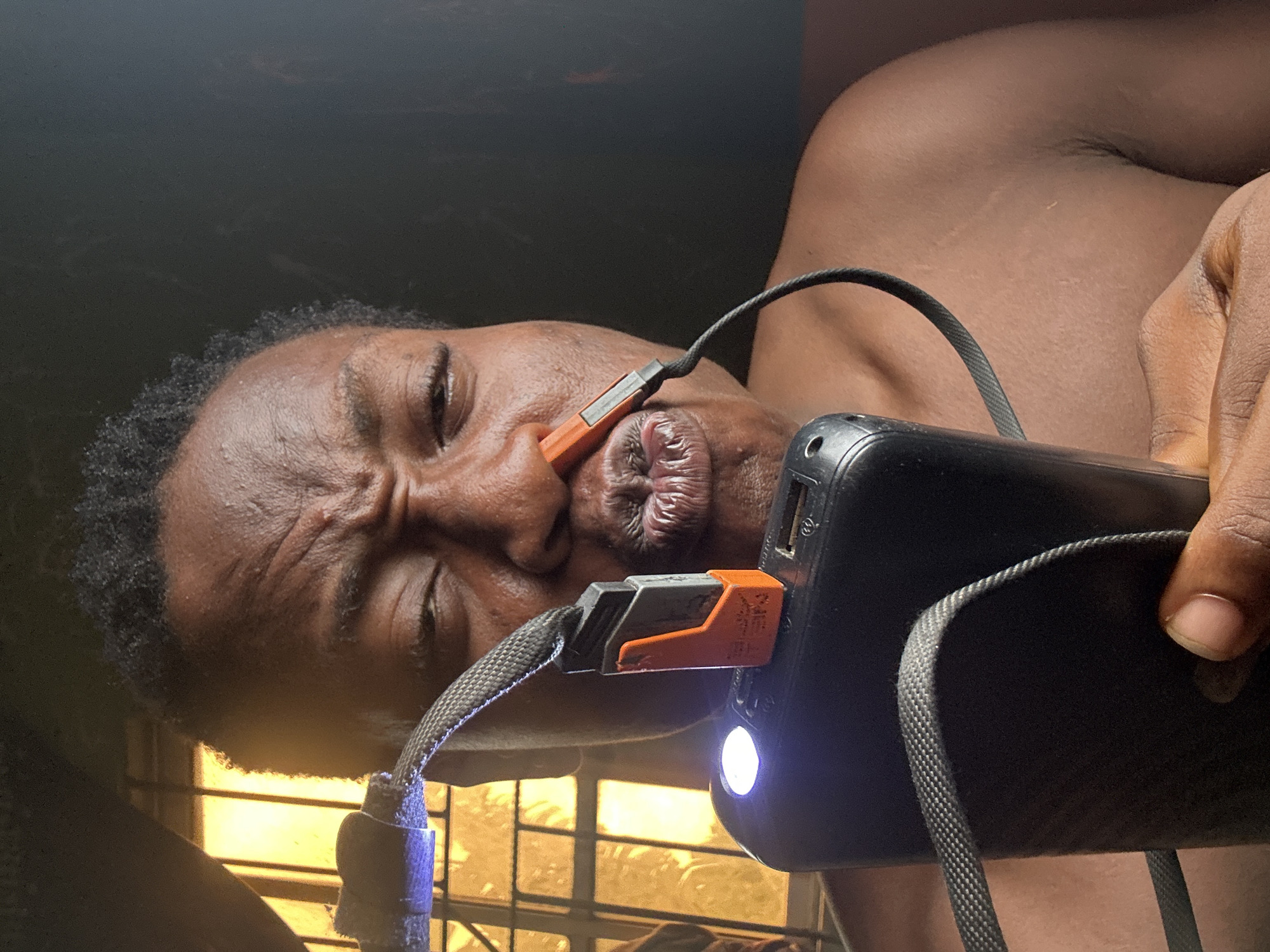 A close-up, portrait-style image captures a person with dark skin and short, dark, curly hair, positioned in a frame that is rotated approximately 90 degrees clockwise. The person's face shows an expression of intense concentration or effort, with eyes partially closed and a furrowed brow. Their lips are distinctly pursed, holding the orange and black connector of a charging cable.
The cable extends from a black portable power bank, which is held by the person's right hand on the right side of the image. The power bank features a bright white LED light that is illuminated. The cable itself has a woven texture and appears to be attached to a dark blue or purple clip or loop near the person's shoulder. Part of the person's bare shoulder and upper arm are visible on the right.
The background is largely dark and indistinct, but a bright, yellowish light source with visible horizontal and vertical bars, resembling a window with blinds or a grate, is present in the lower-left corner, suggesting an indoor setting. The overall lighting is dim, contrasting with the illuminated LED on the power bank. Faint text, "XBET", is visible on the orange connector plugged into the power bank. The image depicts a focused, perhaps unusual, interaction between the person, the charging cable, and the power bank.