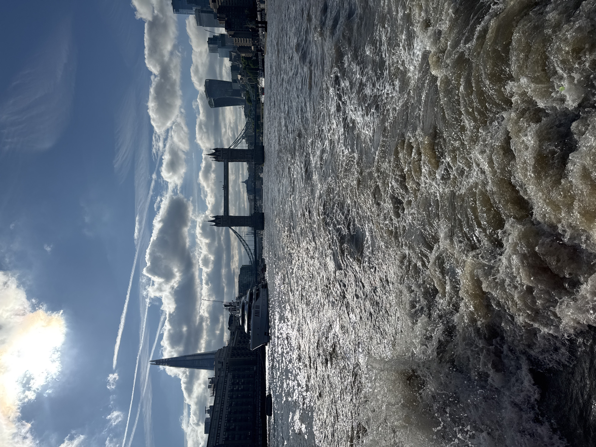This is a vertical, high-angle shot of the River Thames in London, United Kingdom, on a bright, cloudy day. The water is choppy and reflecting the sunlight, creating a sparkling effect. In the background, the iconic Tower Bridge is visible, along with other London skyline buildings, including The Shard. There are also several boats docked along the river. The sky is a mix of blue with white, fluffy clouds, and there are several contrails from airplanes. The overall mood is one of a vibrant, active city day.
