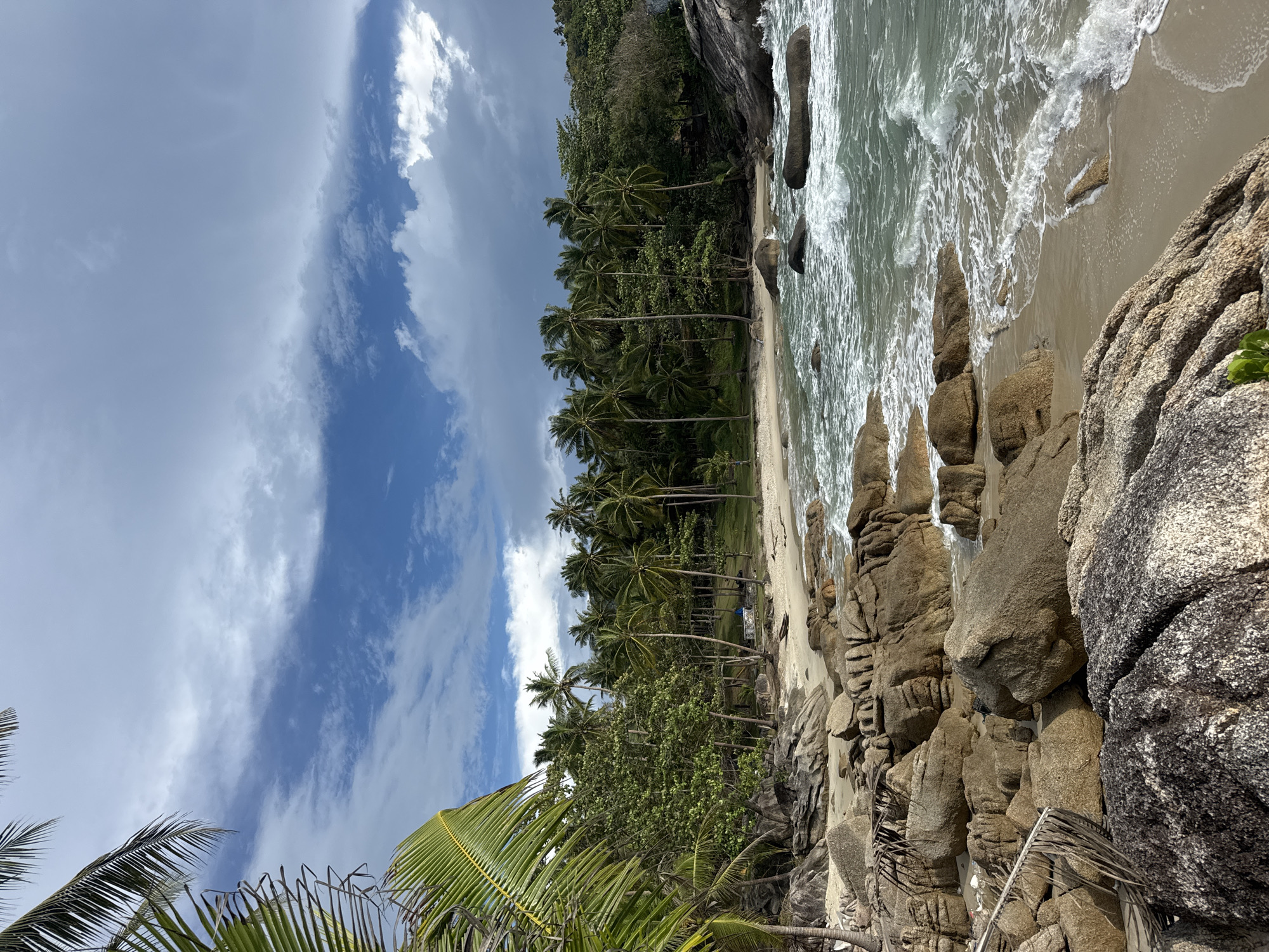The image depicts a beach scene in Baan Thong Nang, Thailand. The view includes a sandy beach with palm trees lining the shore. The sea meets the sand with visible waves, and there are rocky formations along the edge of the beach. The sky above is filled with clouds.