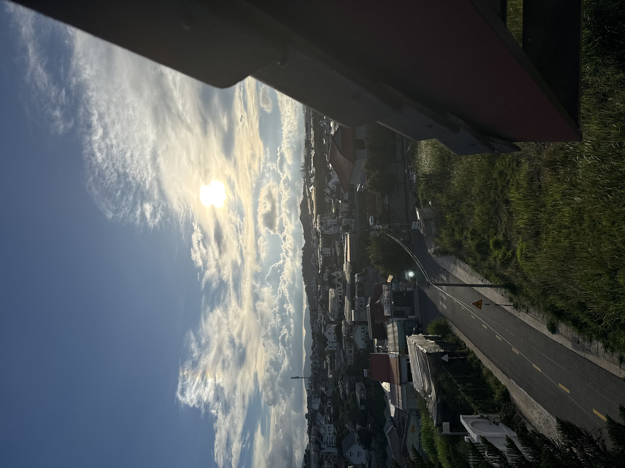 Here's a description of the image:
The image is a high-angle, slightly obscured view over a section of the city of Đà Lạt, Vietnam. The sky occupies a large portion of the scene and is filled with scattered, bright, white clouds against a blue background. The sun is visible and is surrounded by a haze. A gray, possibly metal structure appears at the top of the frame.
Below the sky, the cityscape is visible. It includes numerous buildings, mostly houses with red roofs, densely clustered together. A road cuts through the urban area, with a street light and a traffic sign visible along its edge. Vegetation covers a section of the right side of the image, including grass. In the distance, there are low mountains. The overall lighting suggests it is daytime.