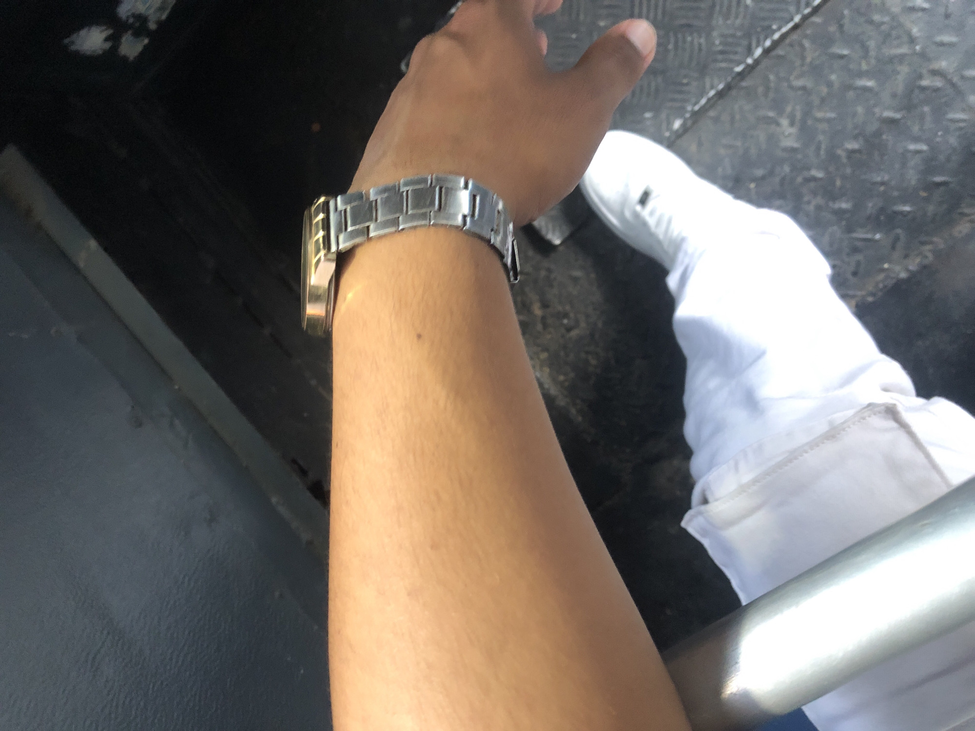 The image shows a person's arm and hand wearing a silver and gold watch. The person is wearing white clothing and shoes. The background consists of gray surfaces, possibly a metal floor or wall. The location is specified as Juan Griego, Venezuela, though this cannot be determined from the image alone. No specific actions or interactions are depicted.
