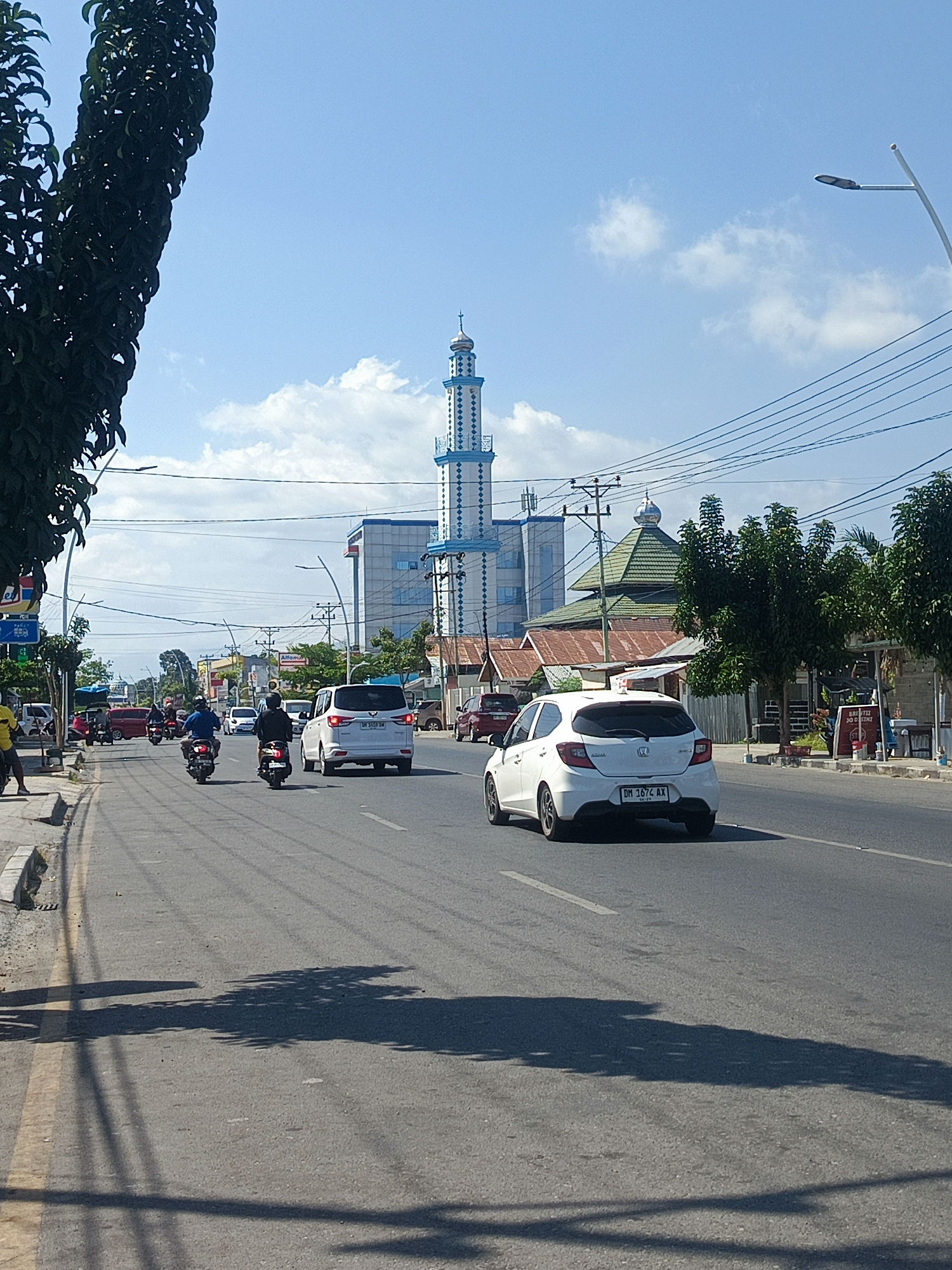 A street scene in Gorontalo, Indonesia, shows a wide asphalt road with a yellow line marking the lane separation. Several vehicles are present, including a white hatchback with license plate DM 1674 AX in the foreground, a white SUV, and multiple motorcycles. A few individuals are visible on foot and on motorcycles. On the right side of the road, there are buildings and trees. Dominating the background is a tall, slender minaret with blue and white accents, a characteristic feature of Islamic architecture. Further back, a building with a green roof and a blue and white structure with a cross on top are visible, suggesting a mix of religious and commercial establishments. The sky is blue with scattered clouds. The image appears to be taken from a moving vehicle, as evidenced by the motion blur and shadows.