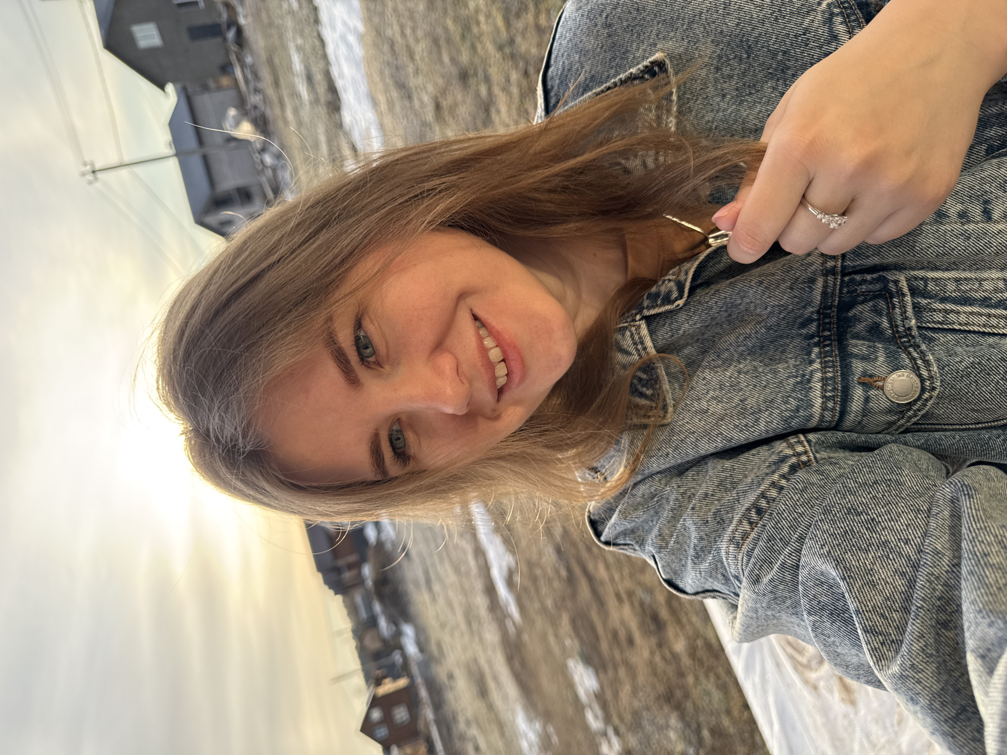 A young woman is seated outside, wearing a denim jacket and a ring on her left hand. She is smiling and looking at the camera. The background shows buildings and fields in the town of Ферма, Russia, with patches of snow visible on the ground. The weather appears to be sunny.