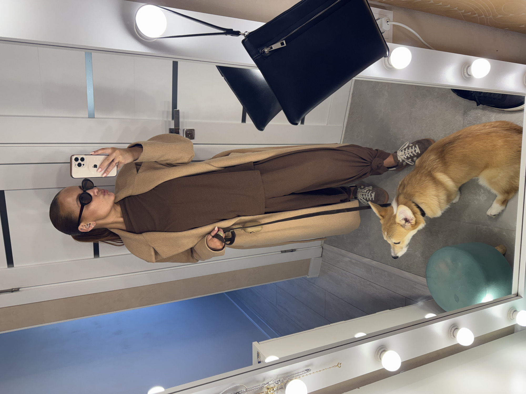 The image shows a woman taking a mirror selfie. She's wearing sunglasses, a brown top and pants, and a tan coat. She holds a smartphone in her right hand to take the picture. A corgi dog stands to her right. A black purse hangs in the upper center of the frame. The mirror is surrounded by lights and appears to be in a dressing room. A small, round green stool sits near the dog. The location of the scene is in the city of Chaikovsky, Russia.