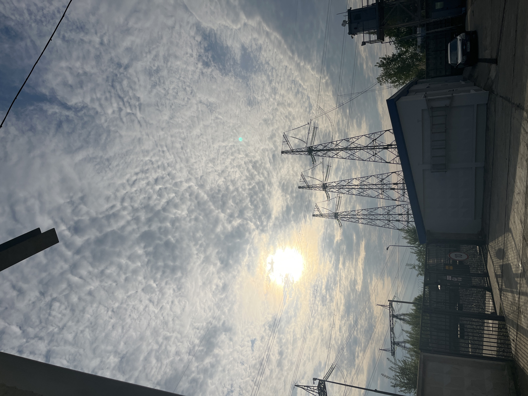 The image, likely taken in Chaikovsky, Russia, presents a low-angle view towards the sky. The sky is filled with altocumulus clouds, with the sun visible through the cloud cover.
Several power line towers are visible, with electrical wires stretching across the frame. A white building with a blue roof line and a ladder is on the right. Behind the building is a black car and what seems to be a gray metal control tower. In front of the building is a dark metal gate and trees. A light pole can be seen too.
The ground appears to be asphalt or concrete. Portions of a gray or concrete structure are visible at the bottom and left edges of the frame.