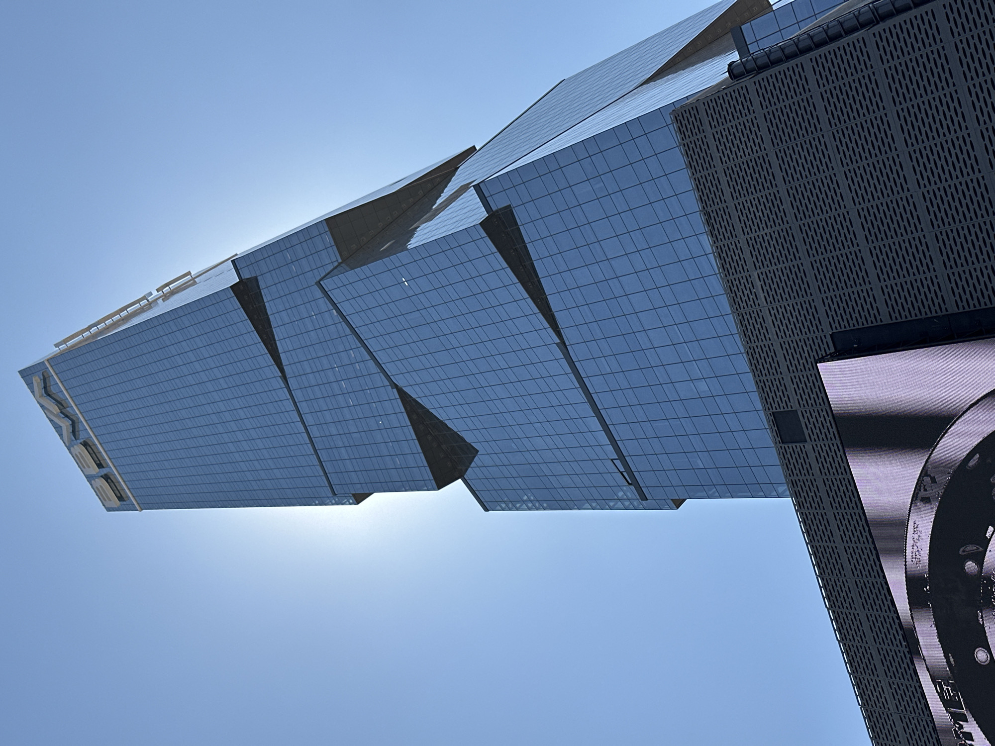 A low-angle upward photograph captures a modern, multi-story skyscraper with a reflective blue glass facade, situated in San Bernardino Tlaxcalancingo, Mexico. The building exhibits a distinctive architectural design characterized by staggered, angular sections and prominent triangular recesses. At its apex, a large, illuminated sign displays the letters "BBVA". The sun, positioned behind the structure, generates a significant backlighting effect and a visible lens flare in the upper left quadrant. The lower portion of the building includes a section with a dark, textured facade composed of perforated panels. Directly below this section, a large, illuminated digital screen is partially visible, displaying an abstract image dominated by a large, circular, mechanical-like component rendered in shades of purple and dark gray. No individuals or discernible actions are depicted in the scene.