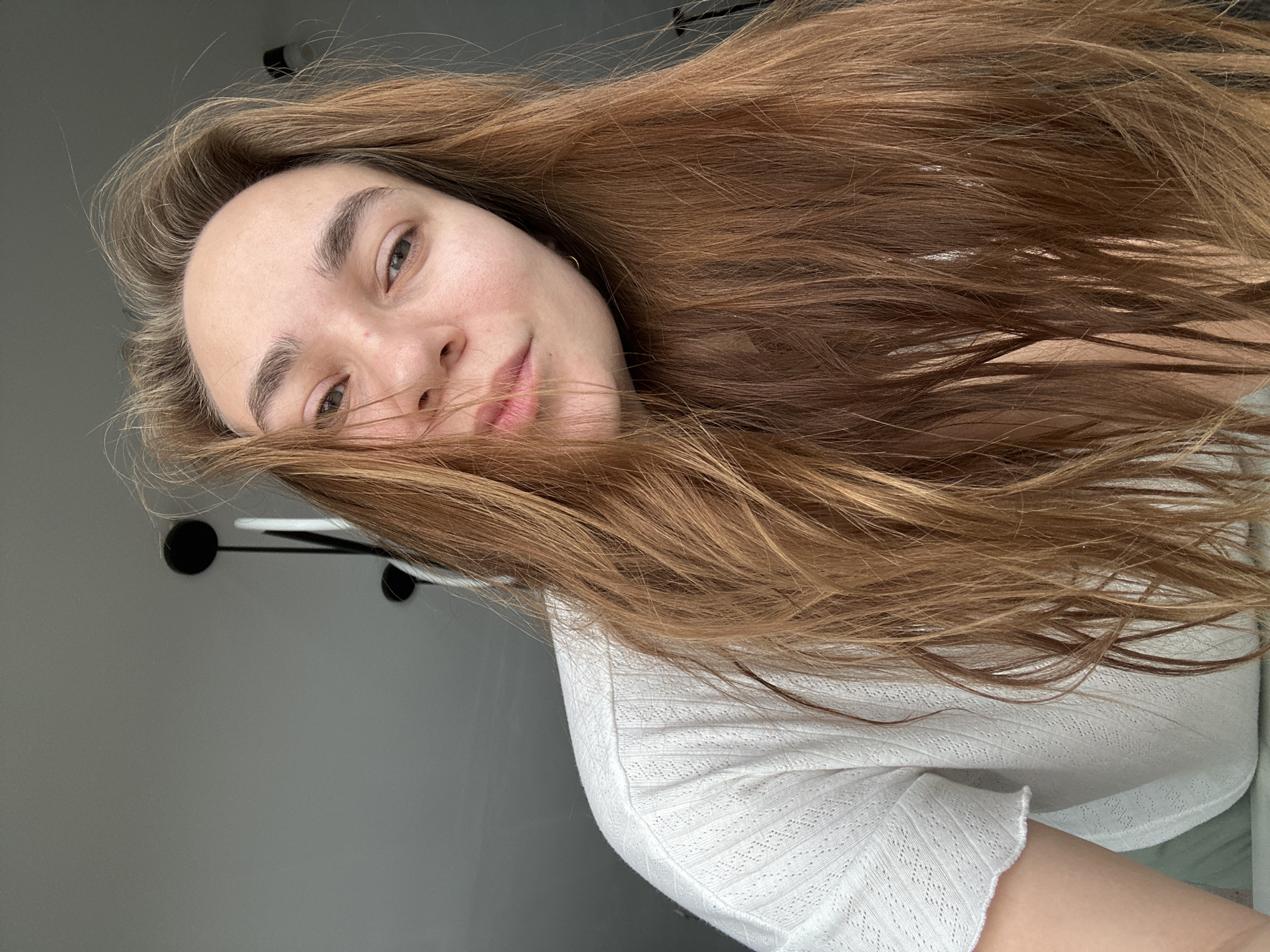 This image depicts a female individual in an indoor setting within Kharnavy, Russia. The subject, positioned centrally, is observed from a high-angle perspective, looking directly towards the camera. She possesses long, light brown hair, appearing somewhat disheveled and framing her face. Her complexion is natural, with light-colored eyes and defined eyebrows. A small, gold-colored hoop earring is visible in her right ear. She is wearing a white or off-white short-sleeved top with a textured, patterned knit. The background consists of a light grey ceiling and wall, with a partial view of a black, multi-stemmed ceiling light fixture visible in the upper right and middle. No other individuals or significant interactions are evident.