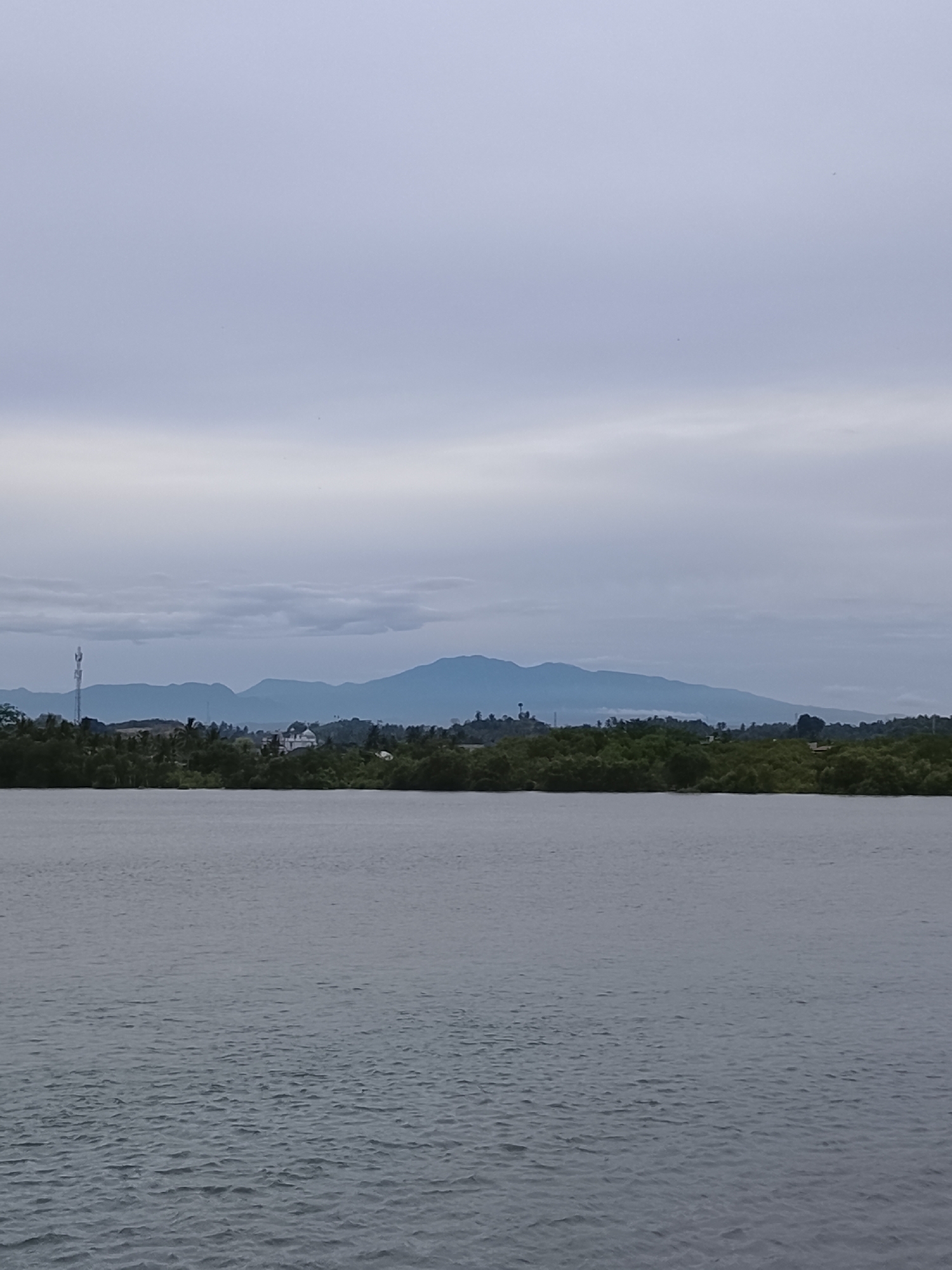 The photograph depicts a landscape scene in Lhokseumawe, Indonesia, dominated by a body of water in the foreground. The water is grey, with visible ripples and small waves across its surface. Behind the water, a strip of land is visible, densely covered with green trees and vegetation, suggesting a shoreline or a forested area. In the distance, a range of mountains can be seen under a cloudy, overcast sky. A cellular tower is visible in the distance, amidst the vegetation. Overall, the scene has a calm, natural aesthetic.