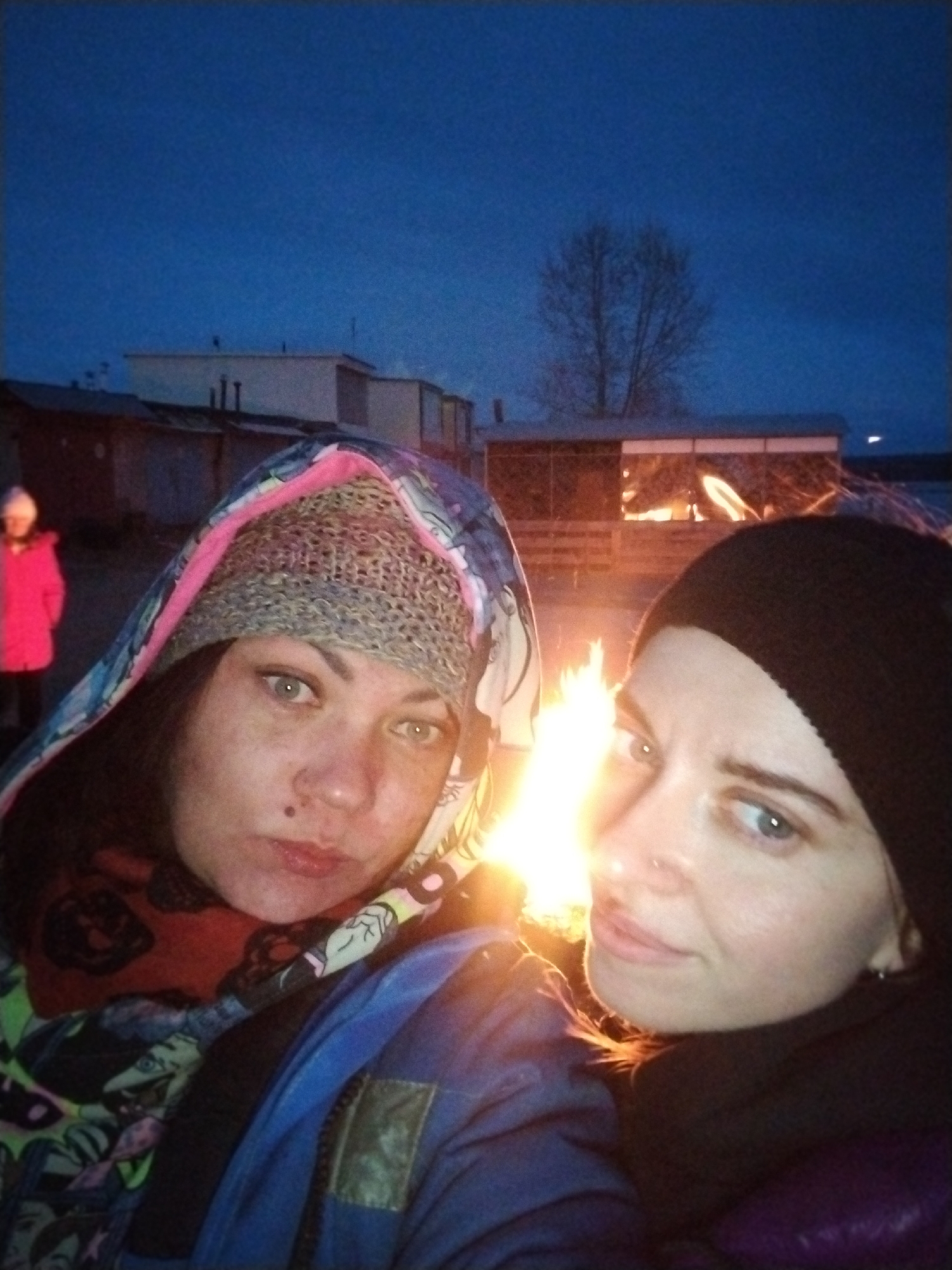 A selfie-style portrait captures two women and a bonfire outdoors at dusk or night in Chaikovsky, Russia. The sky is a deep, dark blue, indicating twilight.
In the foreground, two women pose close to the camera, their faces illuminated by the bright fire. The woman on the left wears a colorful hooded jacket with blue, pink, and green patterns, a gray knitted beanie underneath her hood, and a red patterned scarf. She has dark hair, light blue-green eyes, and a small dark mole above her upper lip. The woman on the right wears a black beanie and a dark jacket, displaying light blue eyes and a nose piercing. Both women look directly at the camera with neutral to slightly relaxed expressions.
A prominent bonfire burns brightly between and slightly behind the two main subjects, casting warm orange and yellow light onto their faces. In the background, to the far left, a third person, likely a child, is visible wearing a bright pink jacket. Behind the fire and people, several low-rise buildings or sheds are silhouetted against the dark sky. One building in the mid-ground has windows reflecting the fire's glow. A bare tree stands silhouetted against the sky in the middle-right background. The heavy clothing worn by the individuals suggests cold weather. There is no visible text in the image.