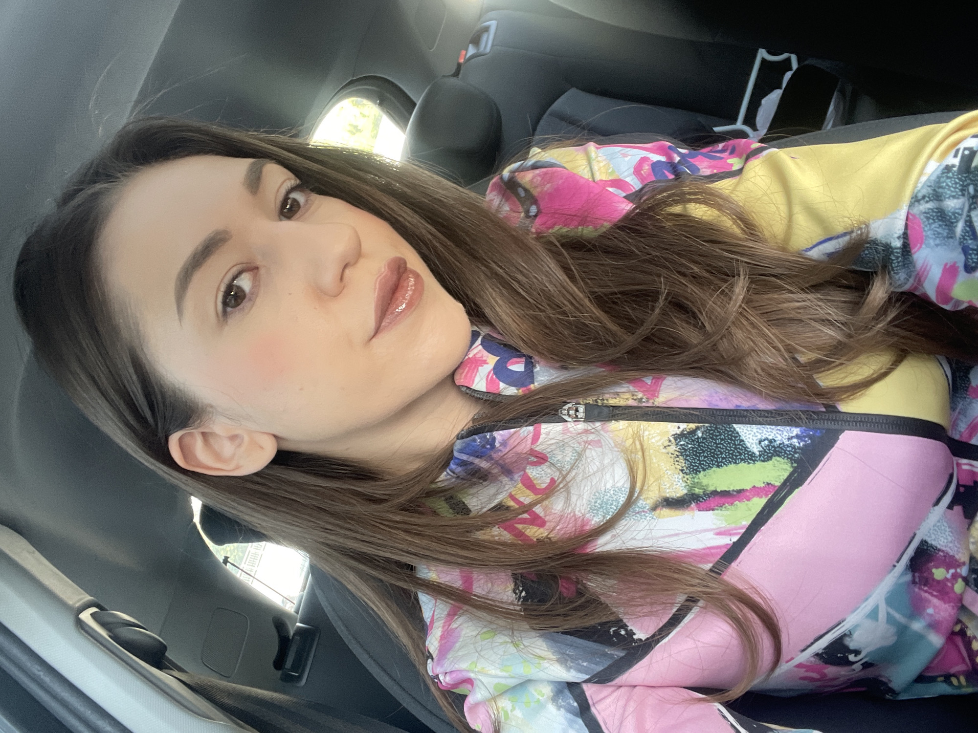 A young woman is depicted in a close-up, slightly tilted portrait, seemingly taken as a selfie inside a car. She has long, straight brown hair, parted on the side, flowing down her shoulders and across her chest. Her face is turned slightly towards the viewer, with a calm, neutral expression and a direct gaze. She wears natural-looking makeup, including defined eyebrows, subtle eye makeup, and glossy, light pink lips. A hint of pink blush is visible on her cheeks.
She is wearing a vibrant, multicolored jacket or top with an abstract, graffiti-style pattern. The garment features bold blocks and splashes of pink, yellow, blue, white, and black. The collar and a zipper with a metallic pull tab are visible at the neckline.
The background suggests the interior of a vehicle, likely a car. A dark gray or black car seat headrest is visible behind her, along with a seatbelt mechanism on the left side of the frame. Through a window in the upper left, blurred greenery and distant structures indicate an outdoor setting during daytime. The lighting is bright and natural. No specific activity beyond posing for the photograph is evident. There is no visible text in the image. The provided context indicates the location is Sofia, Bulgaria.