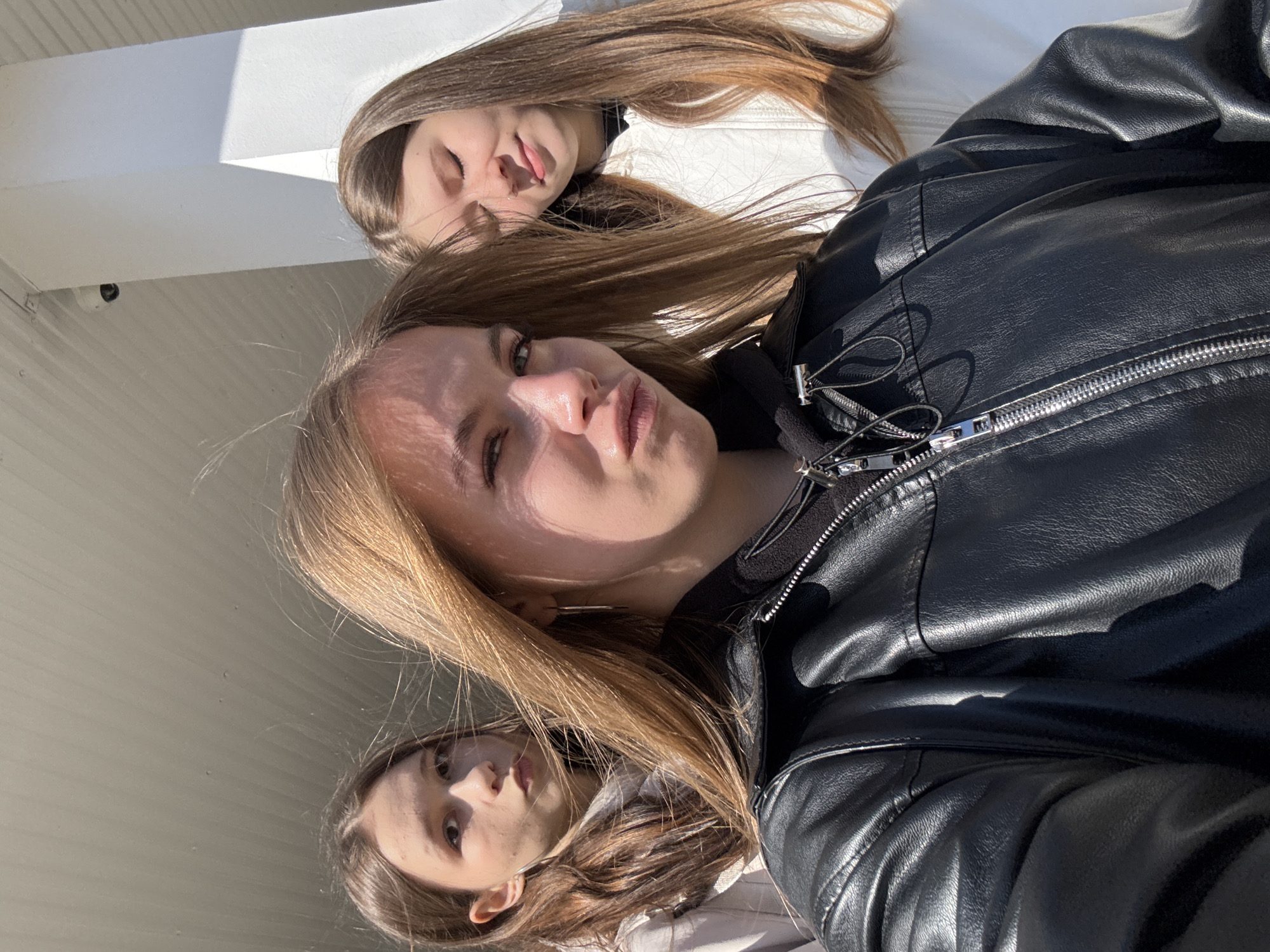 A bright, sunlit group selfie features three young women. The image is oriented vertically, showing the subjects diagonally across the frame.
In the foreground, a young woman with long, straight, light brown hair wears a black leather jacket. Her face, prominent in the center, is illuminated by direct sunlight, casting distinct shadow patterns on her skin. She looks towards the camera with slightly pursed lips.
Behind and slightly above her, another young woman is visible, lying or leaning back. She has long, light brown hair and wears a white top. Her eyes appear closed or squinting, and her tongue is slightly out, conveying a playful expression.
To the lower left, a third young woman's face is partially visible, also looking towards the camera, with long brown hair framing her face.
The background consists of light-colored, possibly white or light grey, architectural structures, suggesting an outdoor or semi-outdoor setting under an overhang or canopy. In the upper left background, a small, white security camera is mounted on the ceiling.
The strong, bright sunlight and sharp shadows indicate a clear, sunny daytime. The overall mood appears casual and relaxed. No visible text is present in the image.
