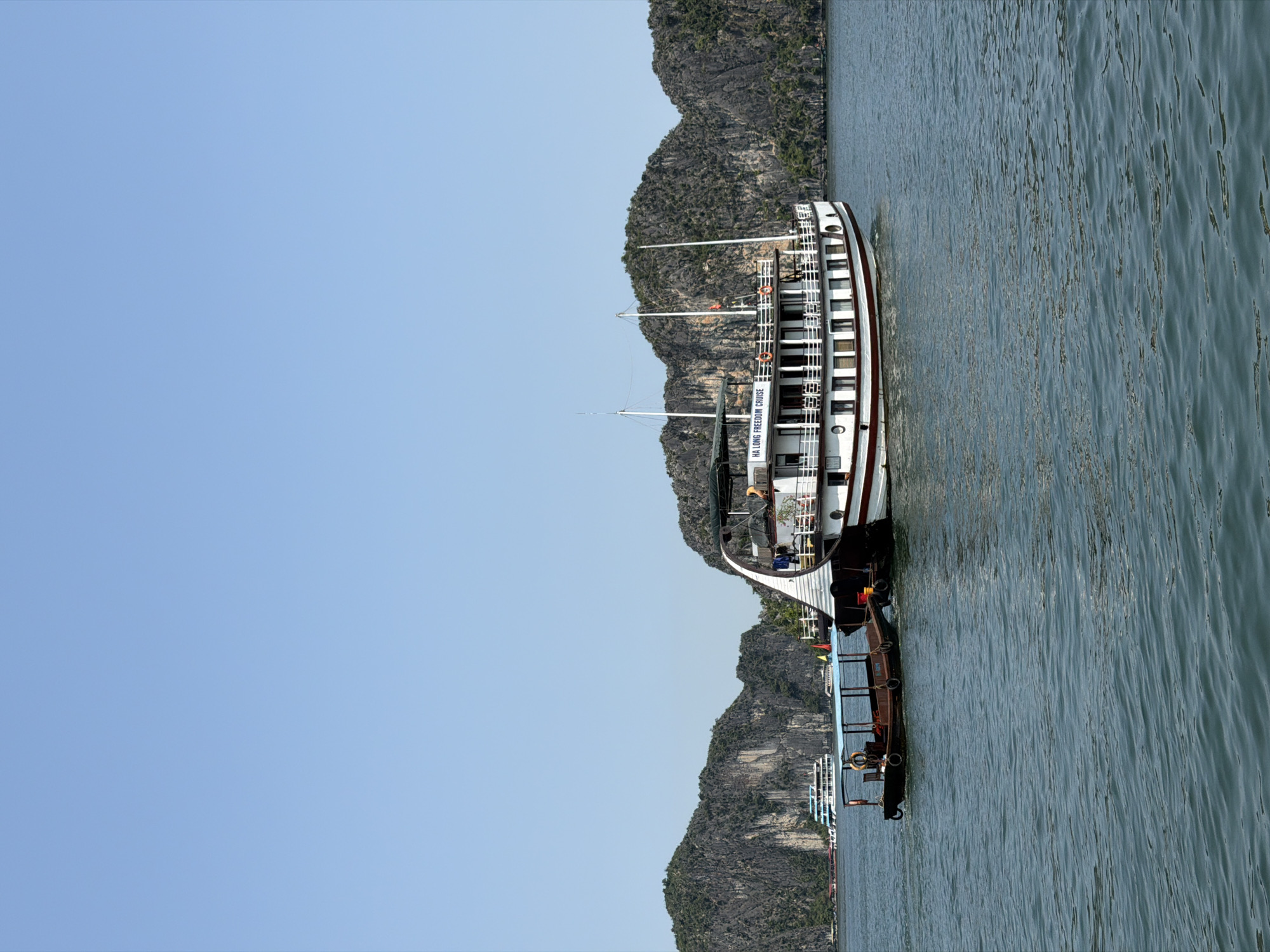 A vertical eye-level shot shows a large boat next to rocky islands in Ha Long Ward, Vietnam.
The boat is white, burgundy, and black, and is on the right of the frame. There are many life vests, and on the side of the boat are the words, "Ha Long Freedom Cruise." Behind the boat is a second, smaller boat. Both are on top of a body of water.
The water takes up the right and bottom of the frame. The sky takes up the left and top of the frame. The islands take up the center of the frame, behind the boats. They are a mix of gray, brown, and green.
