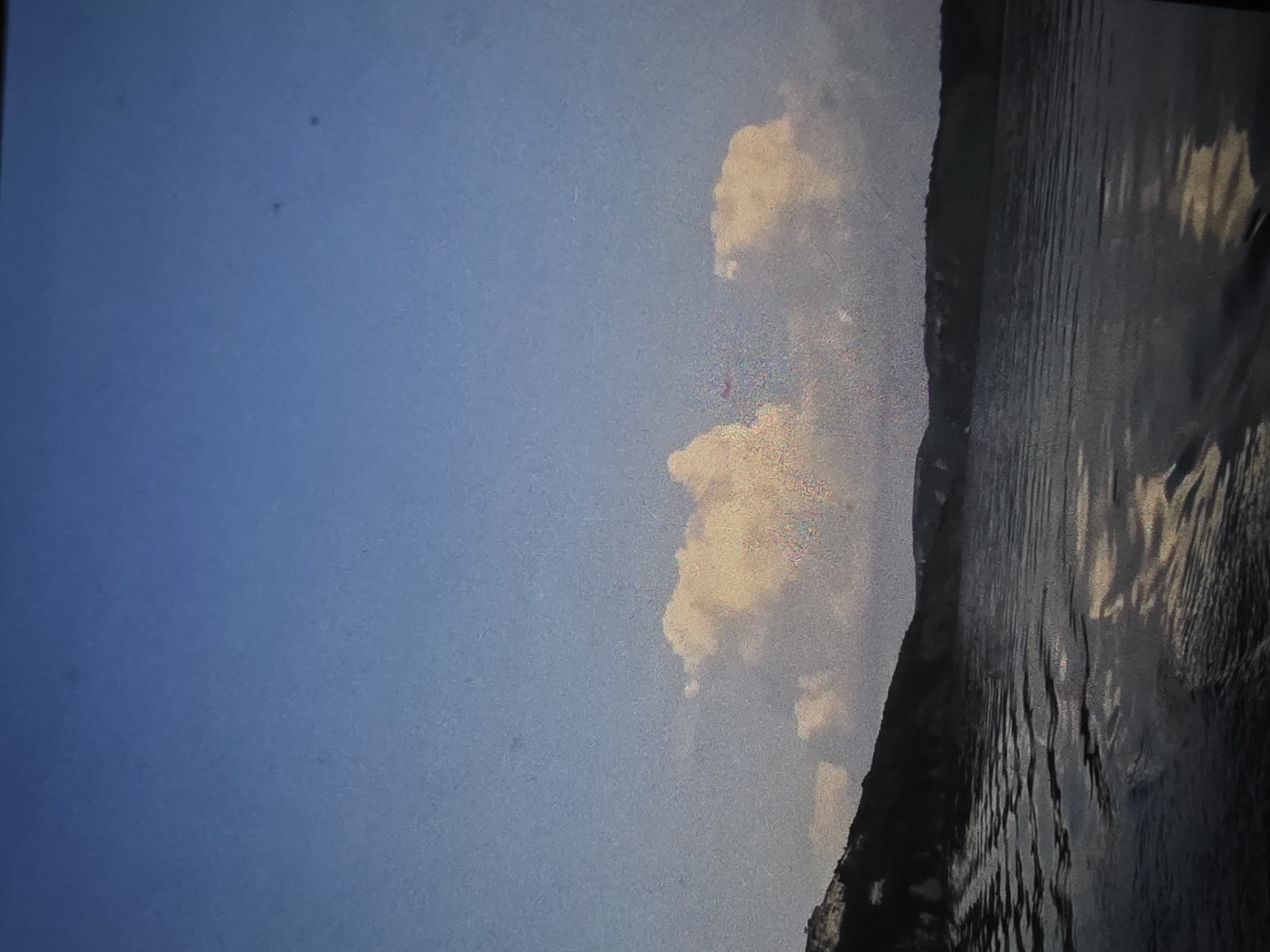 This image depicts a serene landscape with a body of water in the foreground and hills or cliffs in the background. The sky above is a gradient of blue, with some soft, yellowish clouds. The water appears to be calm with gentle ripples reflecting the light from the sky and clouds. The hilly terrain on the horizon is dark, suggesting it's either shaded or covered in vegetation. The scene captures a tranquil natural setting. This image is not taken in Dubno, Ukraine.