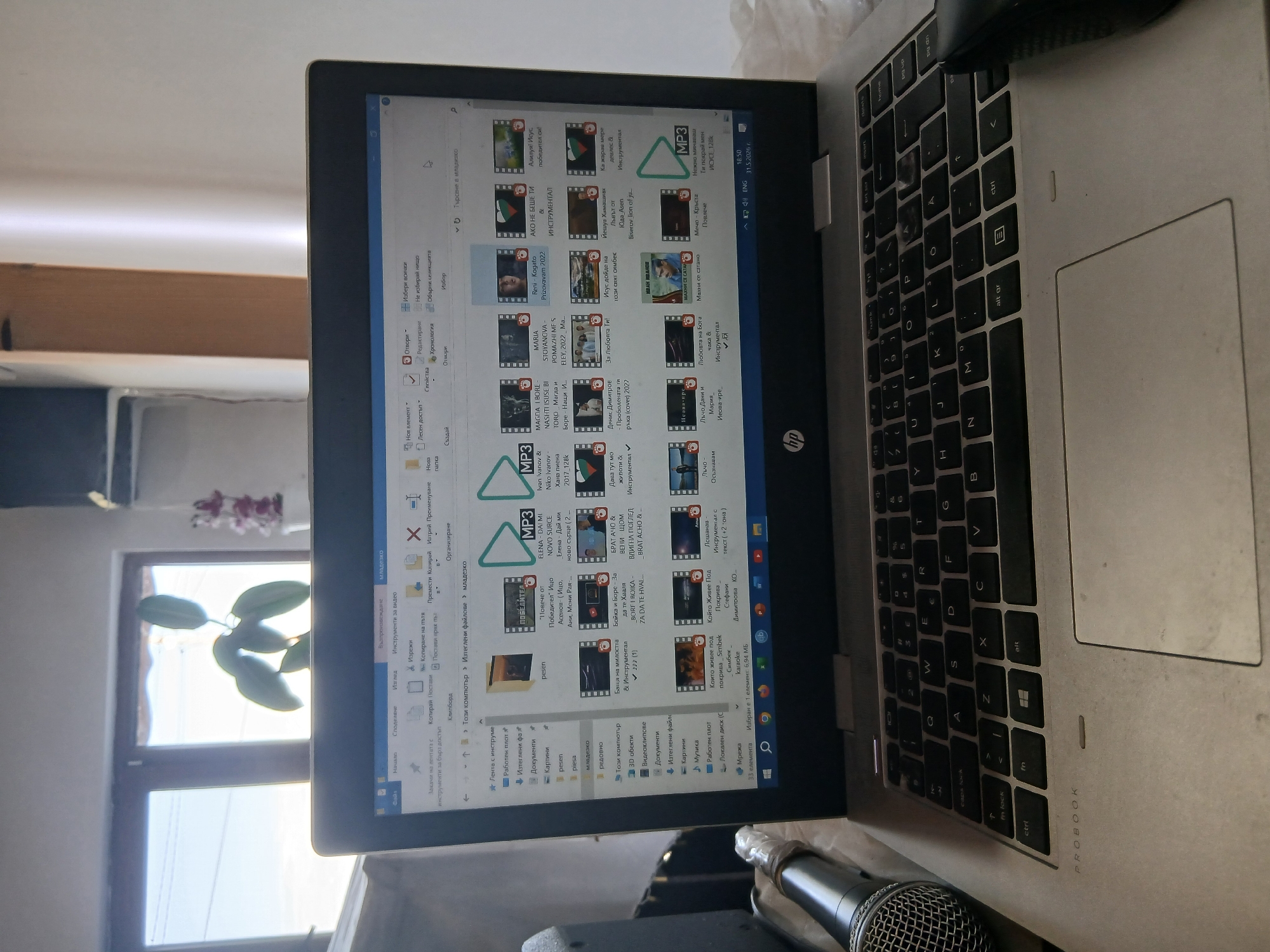 Here is a description of the image:
**Scene:** A close-up shot of an open laptop on a surface, likely indoors.
**Objects:**
* **Laptop:** An HP ProBook, keyboard visible with some wear, showing a Windows file explorer screen filled with various audio and video files. An external mouse is connected to the laptop.
* **Microphone:** A microphone is visible in the lower part of the image.
* **File Explorer:** The screen displays files related to songs and music videos.
* **Window:** A plant is visible in the background behind a window.
**Environment:**
* Appears to be an indoor setting with a window providing natural light, possibly a home or office.
**People:**
* No people are visible.
**Location:**
* The laptop's screen contains Cyrillic text, and the files on the screen suggest a relation to music. Text on the screen indicates "Младежко" (Mladezhko), which could be related to the location or theme of the files. The text on the screen indicates that the laptop is set to Bulgarian.
