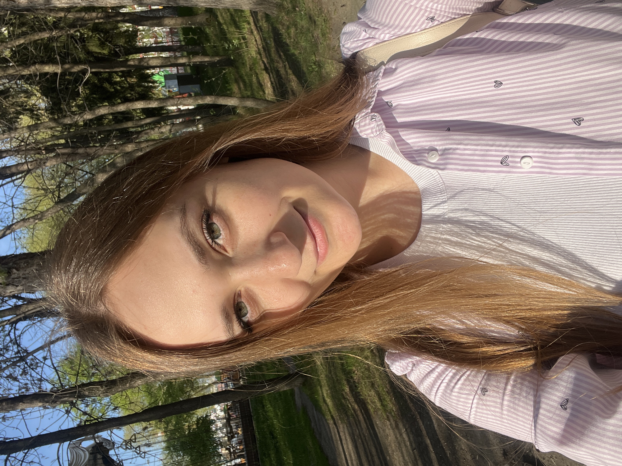 A young woman poses for a selfie outdoors. She has light skin, green eyes, and light brown hair. She is wearing a white ribbed top underneath a striped light purple shirt with a small winged heart pattern. She is standing near trees and a grassy area. A paved path is visible in the background. The location appears to be in a park in Yekaterinburg, Russia, as indicated by the distinctive white pavilion-like structure in the background, which is characteristic of the city's park architecture.