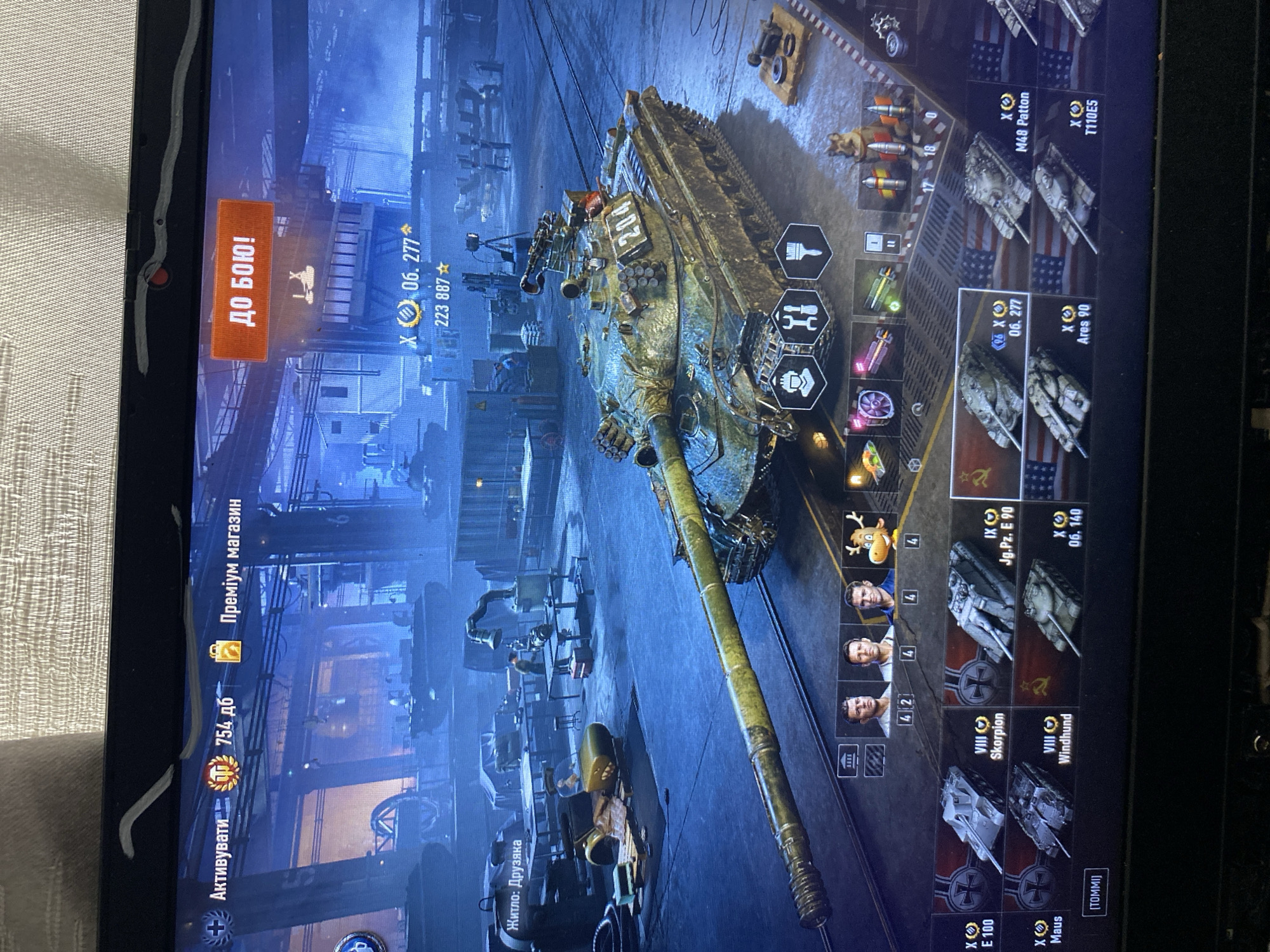 This is a screenshot of a video game, likely World of Tanks, displayed on a laptop screen. The game interface shows a garage environment with a tank as the central subject.
**Environment:** The game displays a virtual garage with industrial elements like scaffolding, workbenches, and other vehicles in the background. The lighting suggests an indoor, possibly dimly lit setting.
**Subject:** The main subject is a heavily armored tank with a long gun barrel. It appears to be a Soviet-era tank based on the markings and design. The tank has the number "204" painted on its side. There are also icons representing crew members and customization options for the tank. In the lower part of the screen, a carousel of other tanks is visible, indicating a selection or comparison interface.
**Interface Elements:**
* "Актививати" (Activate) and "Преміум магазин" (Premium Shop) are visible in the top left.
* "754 дб" indicates some form of in-game currency or points.
* A large orange button with "ДО БОЮ!" (To Battle!) is prominently displayed in the top right, suggesting the player is about to start a game.
* Below the "ДО БОЮ!" button, there is a tank icon and the designation "06. 277" with a number "223 887" below it, likely representing the player's current vehicle and its statistics.
* A text label "Житло: Друзяка" is visible in the upper left, which translates to "Dwelling: Friend" and might be a player nickname or clan tag.
* Various icons represent tank crew members, their skills, and modules.
**Actions:** The scene captures a moment before engaging in a battle, indicated by the "To Battle!" button. The player appears to be in the garage, possibly customizing their tank or selecting a new one.
**People:** There are no identifiable people in the scene other than stylized avatars of crew members within the game's interface.
**Location:** This image is a digital representation within a video game and is not located in the city of Szczecin, Poland, or any real-world location.