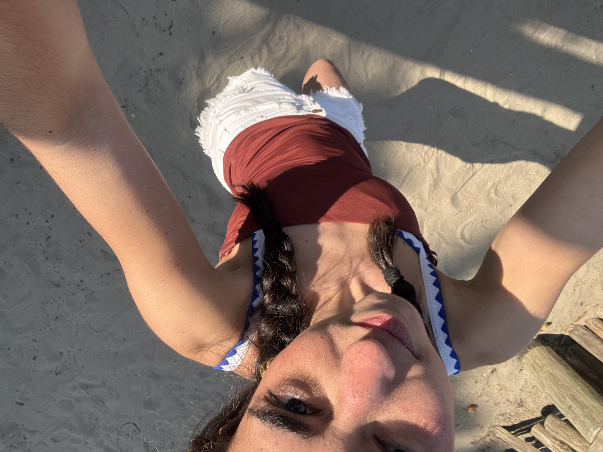 A medium-shot, high-angle photograph shows a person standing on a sandy surface at an unknown location in Unknown, Unknown. The person is wearing a reddish-brown tank top with blue-and-white trim, and white denim shorts. Their hair is styled in braids. The sand has patches of light and shadow, and some wooden objects are visible in the lower right corner of the image. The person holds their arms out to take a selfie-style photo.