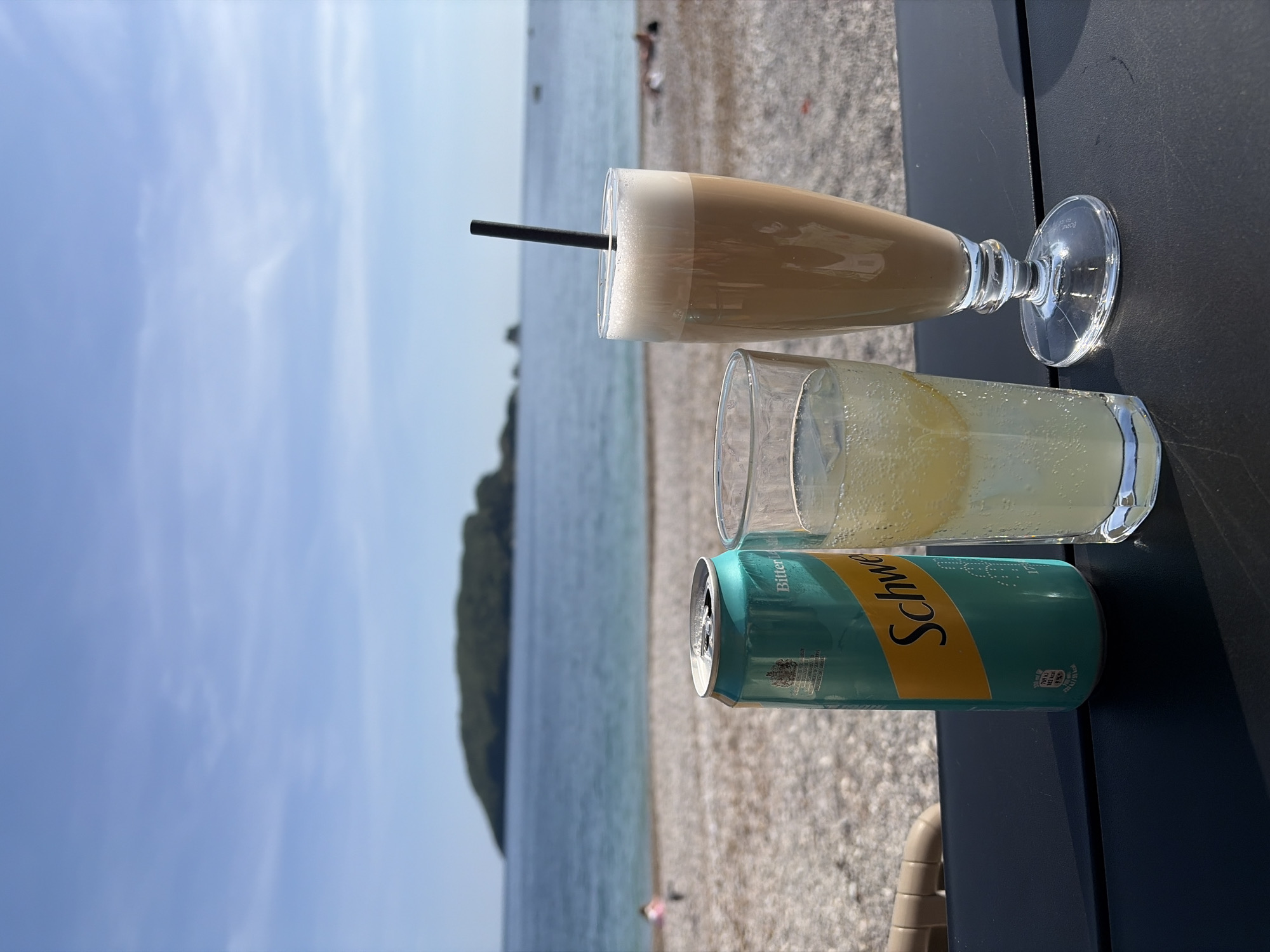 This image depicts a serene beach setting in Budva, Montenegro, on a bright and sunny day. In the foreground, a dark table holds three beverages. On the left, a tall, clear glass contains a layered, light brown drink with a thick white foam top and a black straw, resembling an iced coffee or frappe. To its right, a shorter, clear glass holds a pale yellow, fizzy drink, likely lemonade or a sparkling beverage. Adjacent to this glass is an open, turquoise and yellow aluminum can with the brand name "Schweppes" prominently displayed. Smaller text on the can indicates "Bitter Lemon" and nutritional information, such as "30 kJ / 7 kcal."
The background showcases a pebbly beach leading to a calm, light blue sea. A verdant landmass, possibly a hill or small island, is visible in the distance across the water. The sky above is clear and blue with scattered wispy white clouds. A few blurred figures, likely people, are visible relaxing on the beach in the far background. The scene suggests a relaxed and leisurely time, enjoying refreshments by the sea under clear, sunny weather conditions.