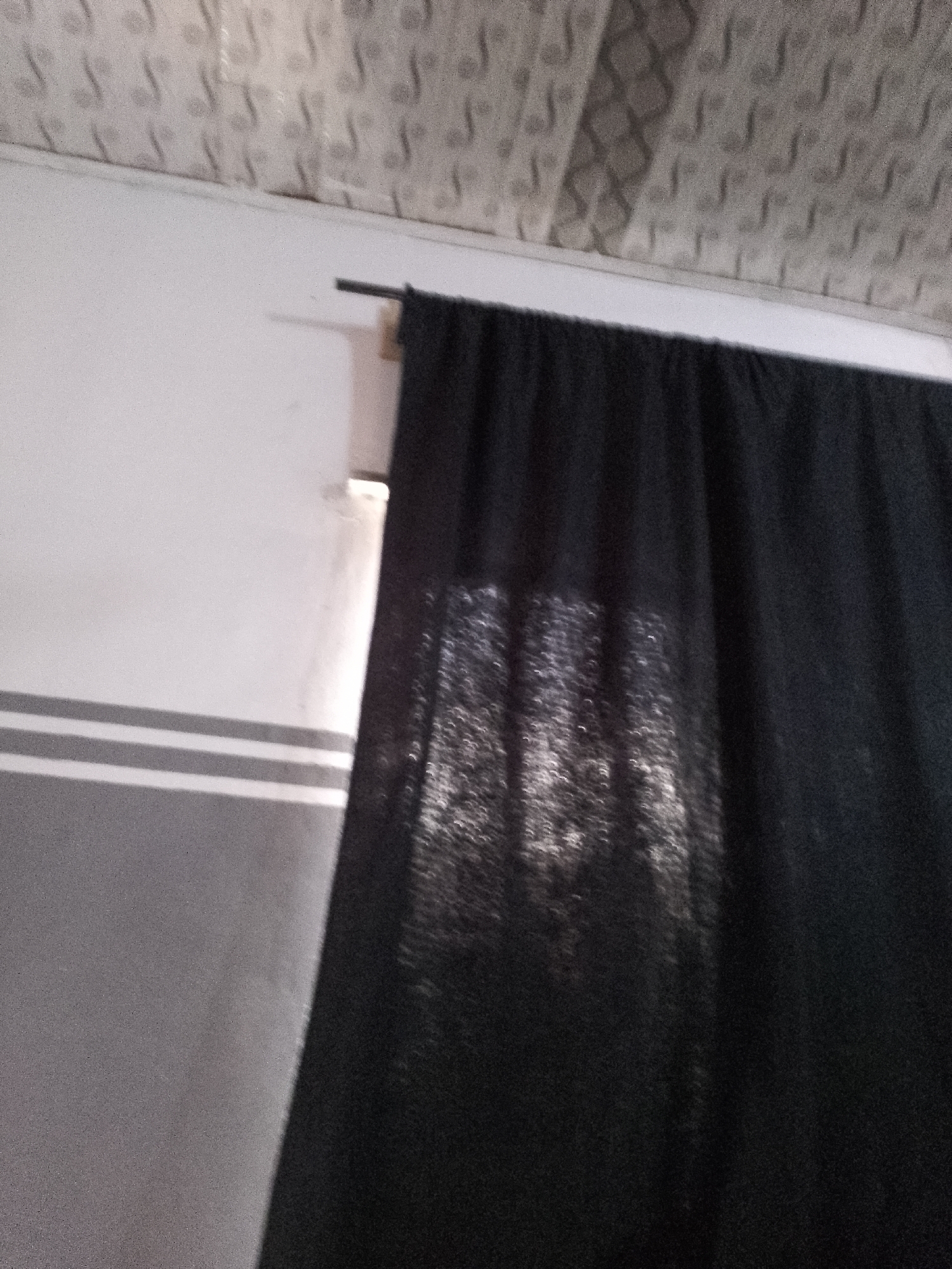 The image shows a black curtain hanging from a rod against a white wall. The curtain is partially transparent, allowing light to filter through and create a pattern of shadows. The ceiling above the wall has a textured, patterned surface. The lower part of the wall has a horizontal stripe design in gray and white. The scene appears to be indoors, in a room with a simple, neutral color scheme. The lighting suggests it might be daytime, with light coming from outside the frame. There are no people or discernible actions in the image.