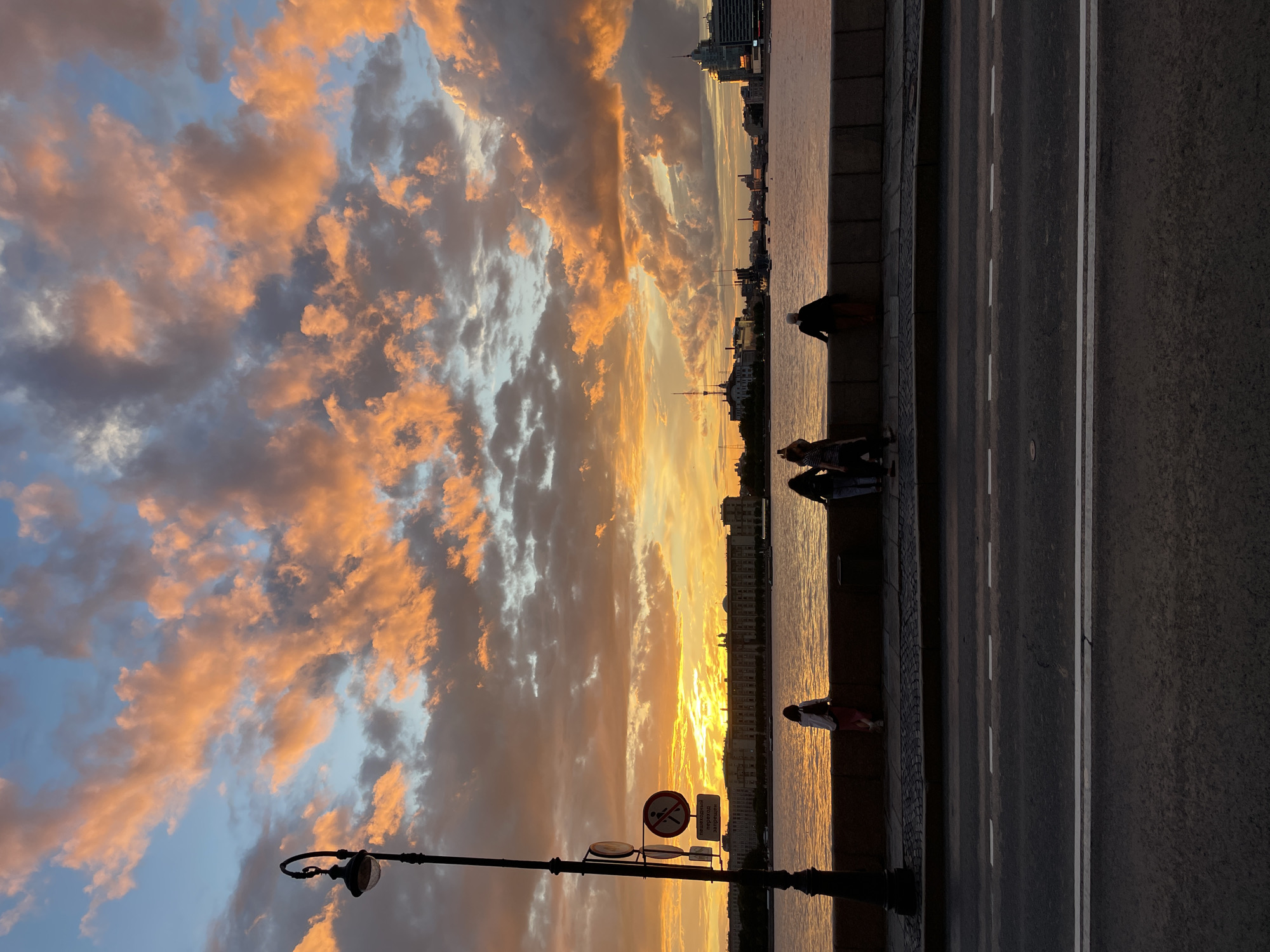 The image shows a waterfront promenade at sunset. The sky is filled with dramatic clouds illuminated by the setting sun, casting an orange glow. A lamppost stands on the left side of the frame, with a circular sign depicting a pedestrian with a diagonal line through it. Several people are visible along the stone embankment, looking out at the water and the cityscape across the river. The buildings across the river appear to be a mix of architectural styles, with some featuring spires and domes. The water is calm, reflecting the colors of the sky. In the foreground, a paved road with white lane markings is visible, suggesting the promenade is adjacent to a street. The overall atmosphere is peaceful and scenic.