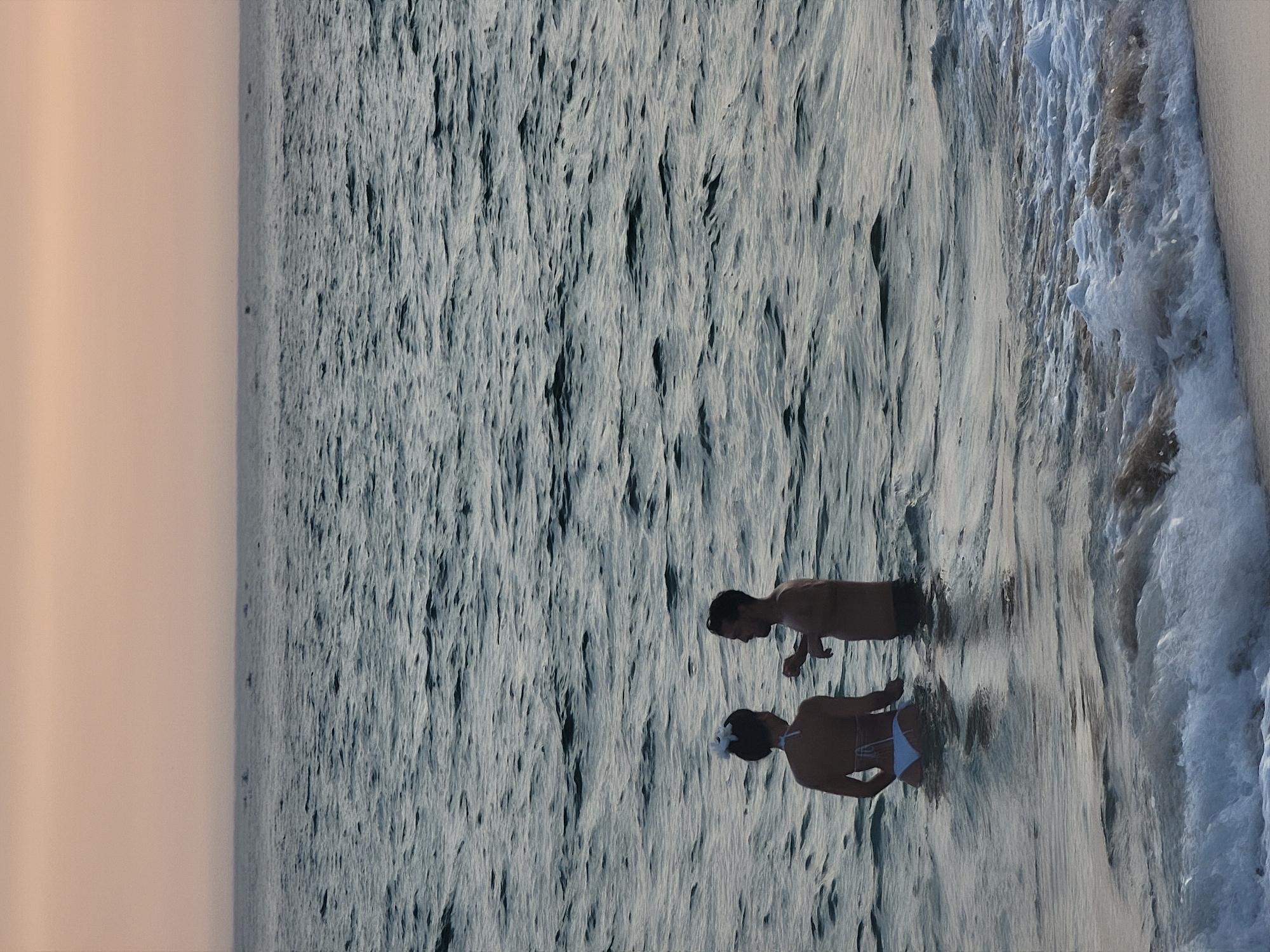 In Quarteira, Portugal, two individuals stand waist-deep in the ocean as waves crash around them. The woman, facing away from the camera, wears a white bikini and has a white flower in her hair. The man, facing the woman, wears dark swim trunks and gestures with his right hand. The sky above is a soft, muted orange, suggesting either sunrise or sunset, and the water is a deep, greyish-blue with rippling waves. The scene captures a moment of quiet interaction between the two people in the serene, natural environment of the sea.