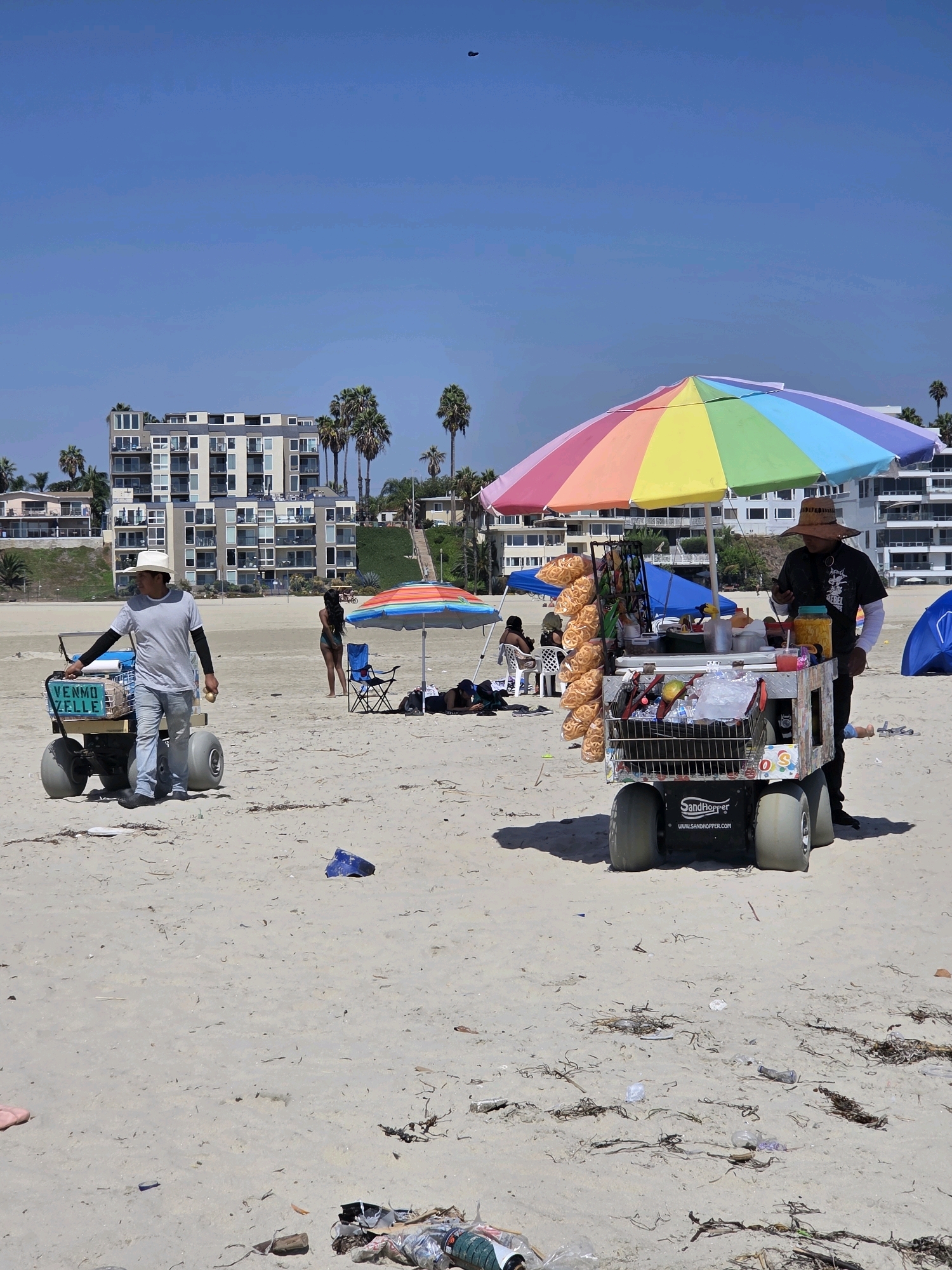 The image shows a sunny day at a beach in Long Beach, California. A man wearing a white hat and a gray shirt is pushing a cart on the sand. The cart has large wheels and a sign on the side that says "VENMO ZELLE". Another man, wearing a straw hat and a dark t-shirt, is standing behind a colorful umbrella-covered cart that is selling snacks. The cart is stocked with bags of chips, drinks, and other food items. In the background, there are several large apartment buildings, palm trees, and other beachgoers. The sky is clear and blue. The sand is light brown and scattered with debris.