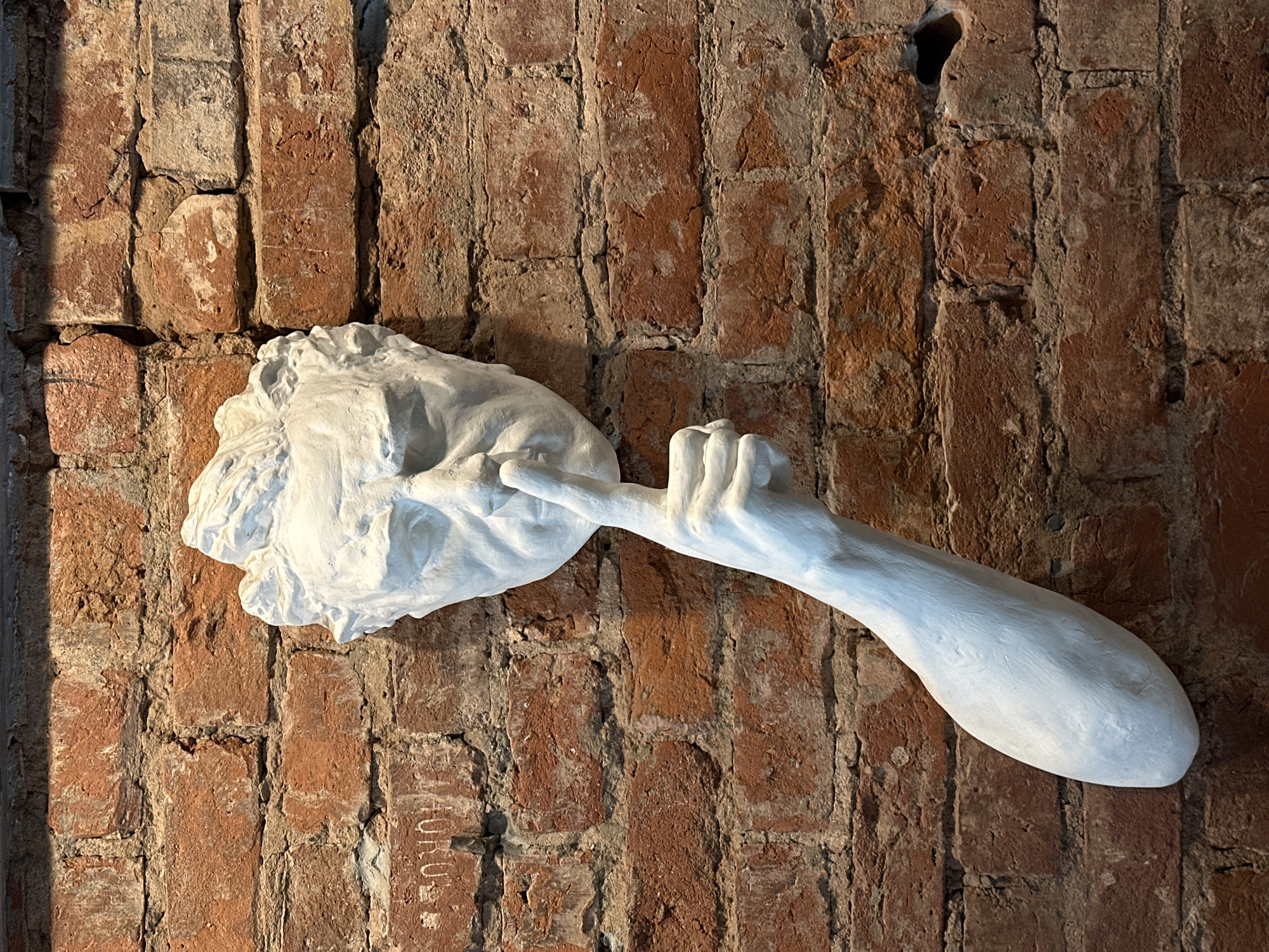 The image displays a white sculpture of a head and an arm attached to a brick wall. The sculpture depicts a man with his finger held to his lips, in a gesture of silence. The wall is made of red bricks, some of which are worn and have visible mortar. There are some inscriptions on the wall, partially visible. The sculpture appears to be art displayed on the wall. The location is identified as Moscow, Russia.