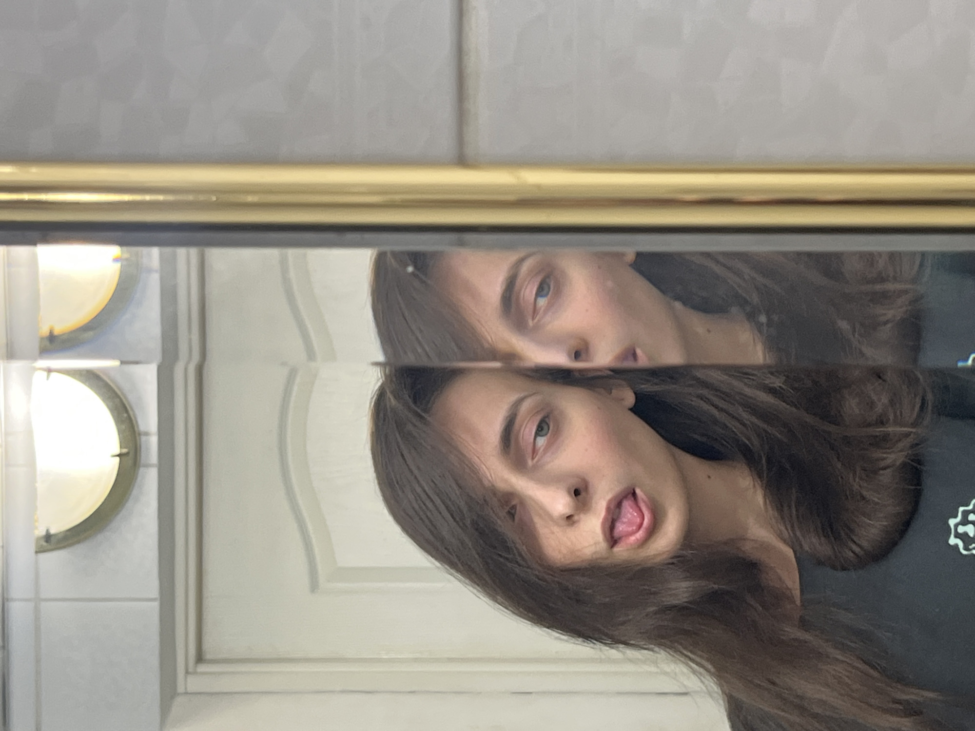 A person is reflected in a bathroom mirror. The person has long, brown hair and is sticking their tongue out. A gold frame is visible at the top of the mirror. The bathroom has white walls and a light fixture. There is no information about the location in Krasnoyarsk, Russia in the picture.