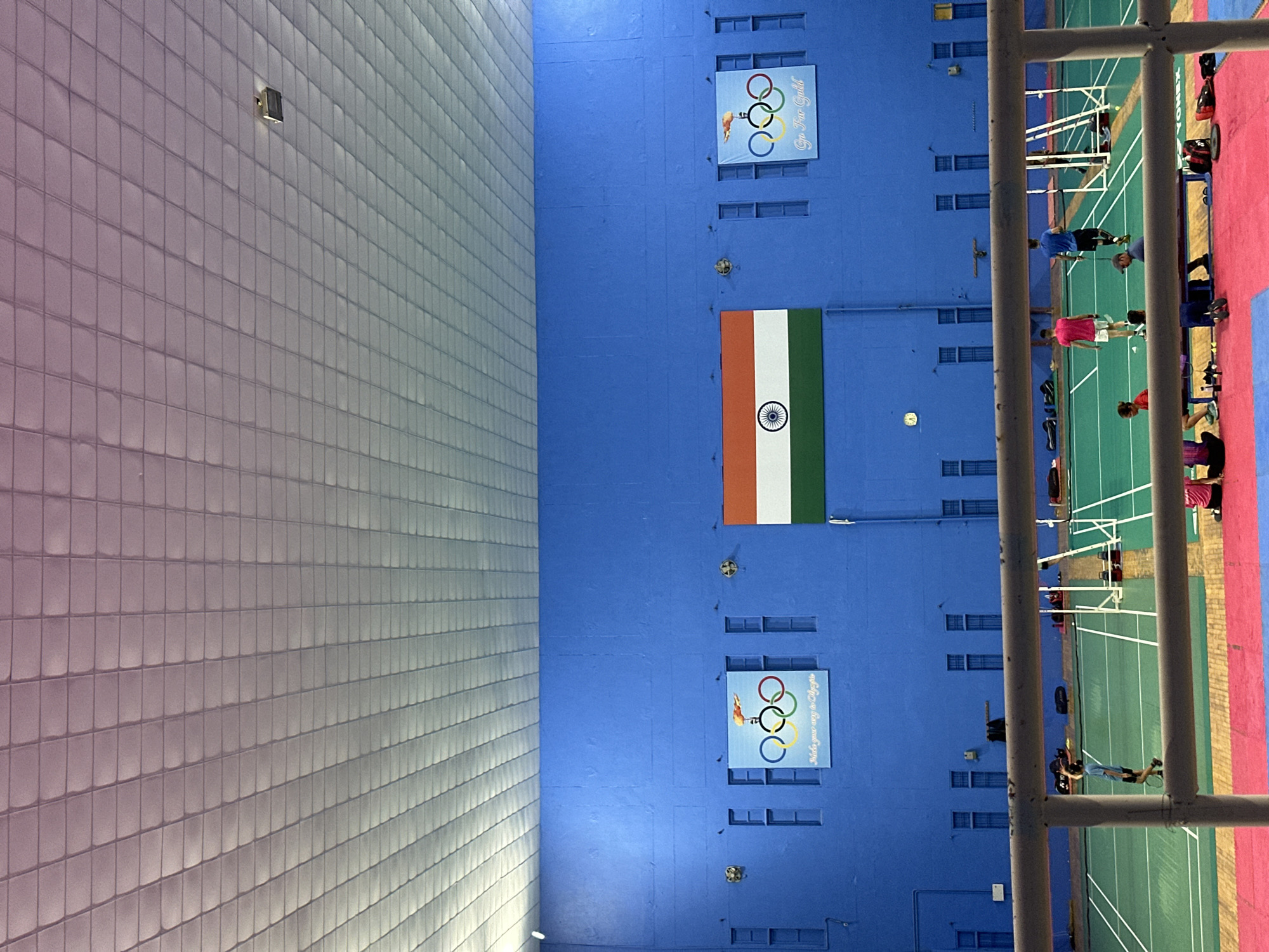 An indoor sports facility with a blue wall features Olympic rings with a tagline and the Indian flag. The ceiling is a white, grid-like structure with lights. Below the wall, a green badminton court is visible with white lines marking the boundaries. Several people are on or near the court, some are playing badminton, and others are sitting or standing around. A metal railing runs across the foreground. The overall environment suggests a training or practice session for badminton players.