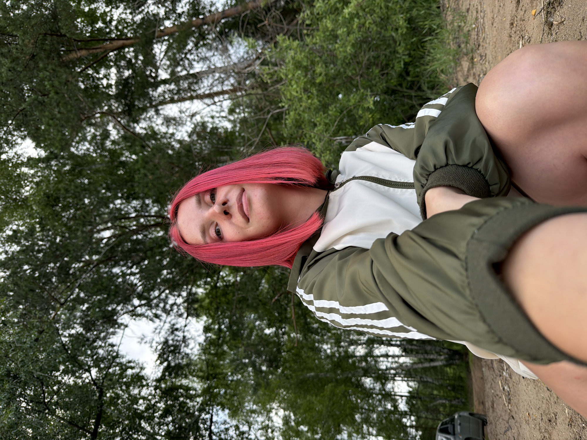 The media file is a close-up, vertical, selfie-style shot of a young person with vibrant pink hair. They are wearing a green and white jacket with white stripes on the sleeves. The person is looking towards the camera with a slight smile. The background consists of a blurred forest with green trees and some sky visible. The ground in the foreground appears to be sandy or dirt, with some grass. A small portion of a car can be seen in the lower right background. The lighting suggests it is daytime, possibly overcast. The overall mood appears casual and relaxed, as if the person is outdoors. There is no visible text in the image. The location context is Chaikovsky, Russia.