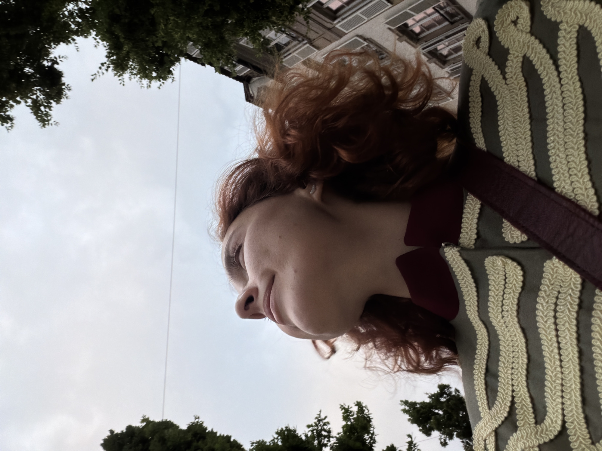 A low-angle, close-up shot captures a young woman with auburn, curly hair looking upwards. She is wearing a dark red collar and a green jacket adorned with intricate gold braiding. A dark red strap is visible across her chest. The background shows a cloudy sky with a power line stretching horizontally. To the right, a building with shuttered windows and lush green trees framing the scene suggests an urban outdoor environment, likely in the city of Strasbourg, France.