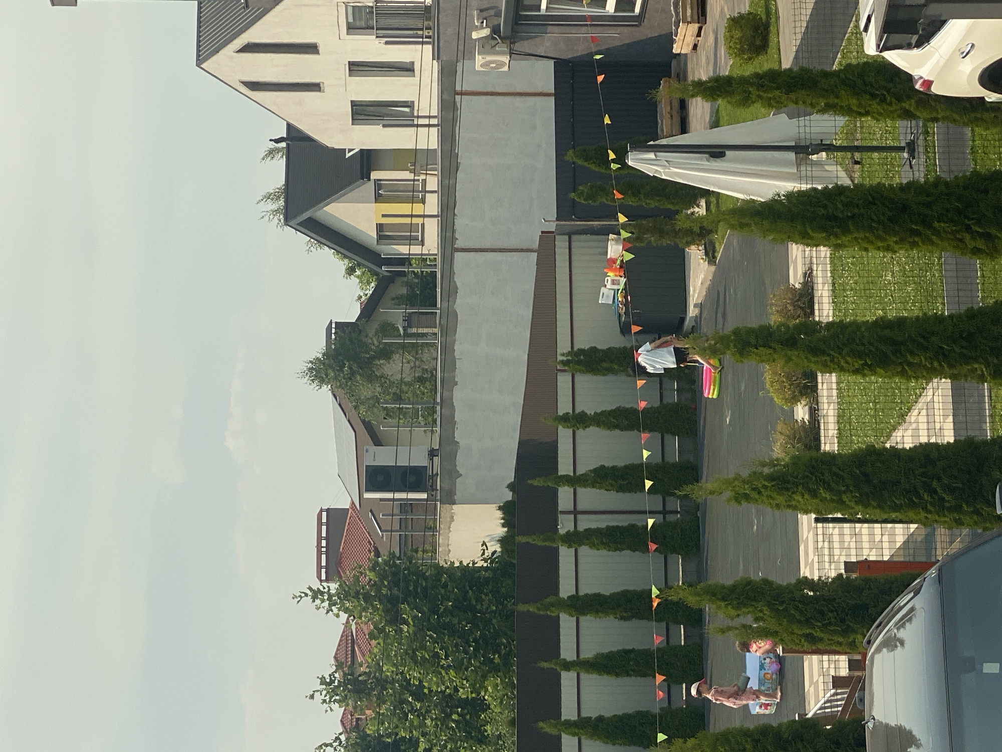 The image shows a residential street scene in Novi Petrivtsi, Ukraine. The foreground features a row of manicured evergreen trees and a portion of a car. Beyond the trees, a paved driveway leads to a multi-story building with a modern architectural design. A white umbrella is set up on the grass next to the driveway. A string of colorful triangular flags is strung across the scene, suggesting a festive atmosphere or an event.
In the mid-ground, two children are visible near a low table with toys, indicating a playful activity. One child is seated and appears to be engaged with the toys, while the other stands nearby, possibly playing as well. Further in the background, more residential buildings are visible, some with red-tiled roofs. The sky is bright and appears to be a clear day, suggesting it is daytime. The overall setting appears to be a well-maintained suburban or semi-rural residential area. No visible text is present in the image.