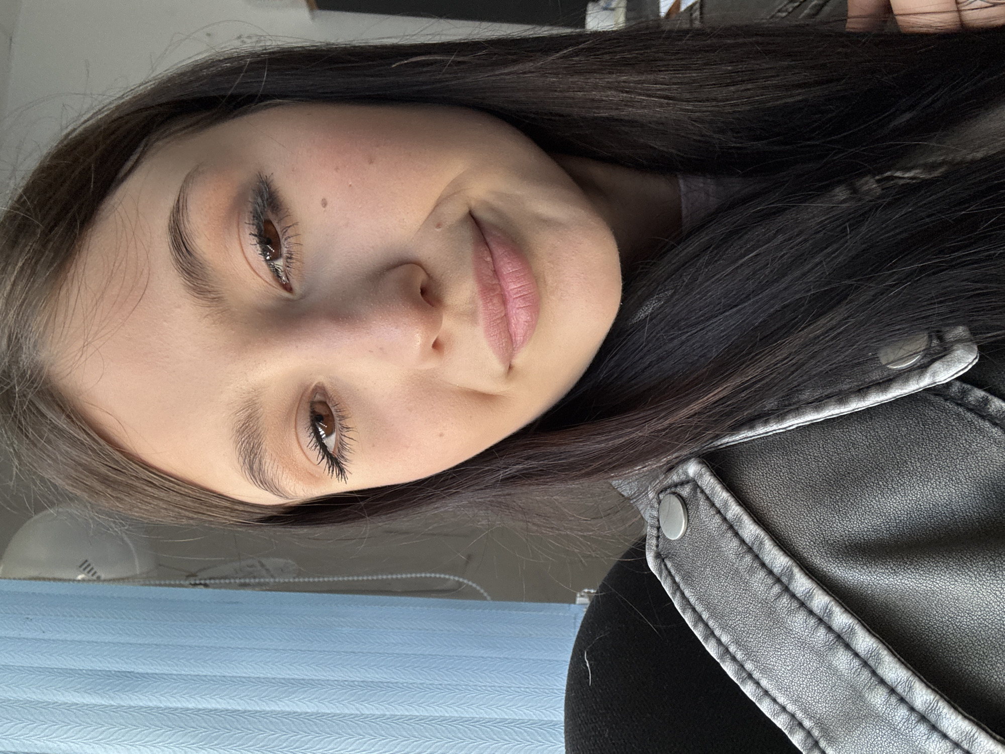 A vertical portrait features a young woman with long, dark brown hair, looking directly at the camera with a subtle, gentle smile. She appears to be in her late teens or early twenties, with her head slightly tilted. Her brown eyes are accentuated with mascara and a light peach or brown eyeshadow, and her lips have a soft pink hue. Her skin shows a few small moles.
She is dressed in a dark, possibly black, leather-style jacket with visible stitching and silver snap buttons on the lapel/shoulder area, worn over a dark undershirt. The background is softly blurred, indicating an indoor setting. To her left, light blue textured horizontal blinds are visible, suggesting a window and a source of natural light. A faint, out-of-focus white object can be seen above the blinds. The overall lighting is soft, consistent with daylight. The scene, located in Chaikovsky, Russia, suggests a casual and relaxed atmosphere. No text is visible in the image.