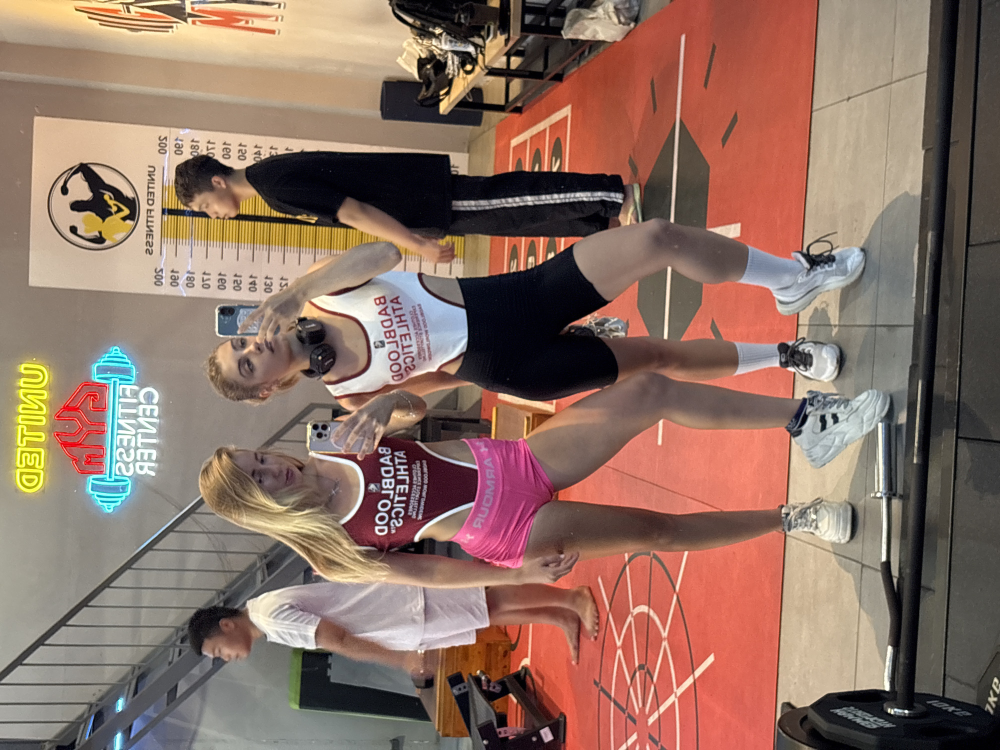Here is a description of the image:
**Scene:** Indoor fitness center.
**People:** Three individuals are captured. One individual is a young woman with blonde hair wearing a burgundy and white top, pink shorts, and white sneakers. She appears to be taking a selfie. Another woman with light brown hair is behind her. She is wearing a white and black top, black shorts, white socks, and white sneakers. She also appears to be taking a selfie. A male individual is standing further back, wearing a black shirt and black striped pants. Another man is standing in the background wearing a white shirt and white shorts.
**Objects:** Gym equipment including weights and bars are visible. There is a height measurement chart on the wall in the background and a neon sign that says "United Fitness Center".
**Actions:** Individuals are standing and presumably taking photos.
**Environment:** The scene appears to be inside of a fitness facility with a workout area marked on the floor.