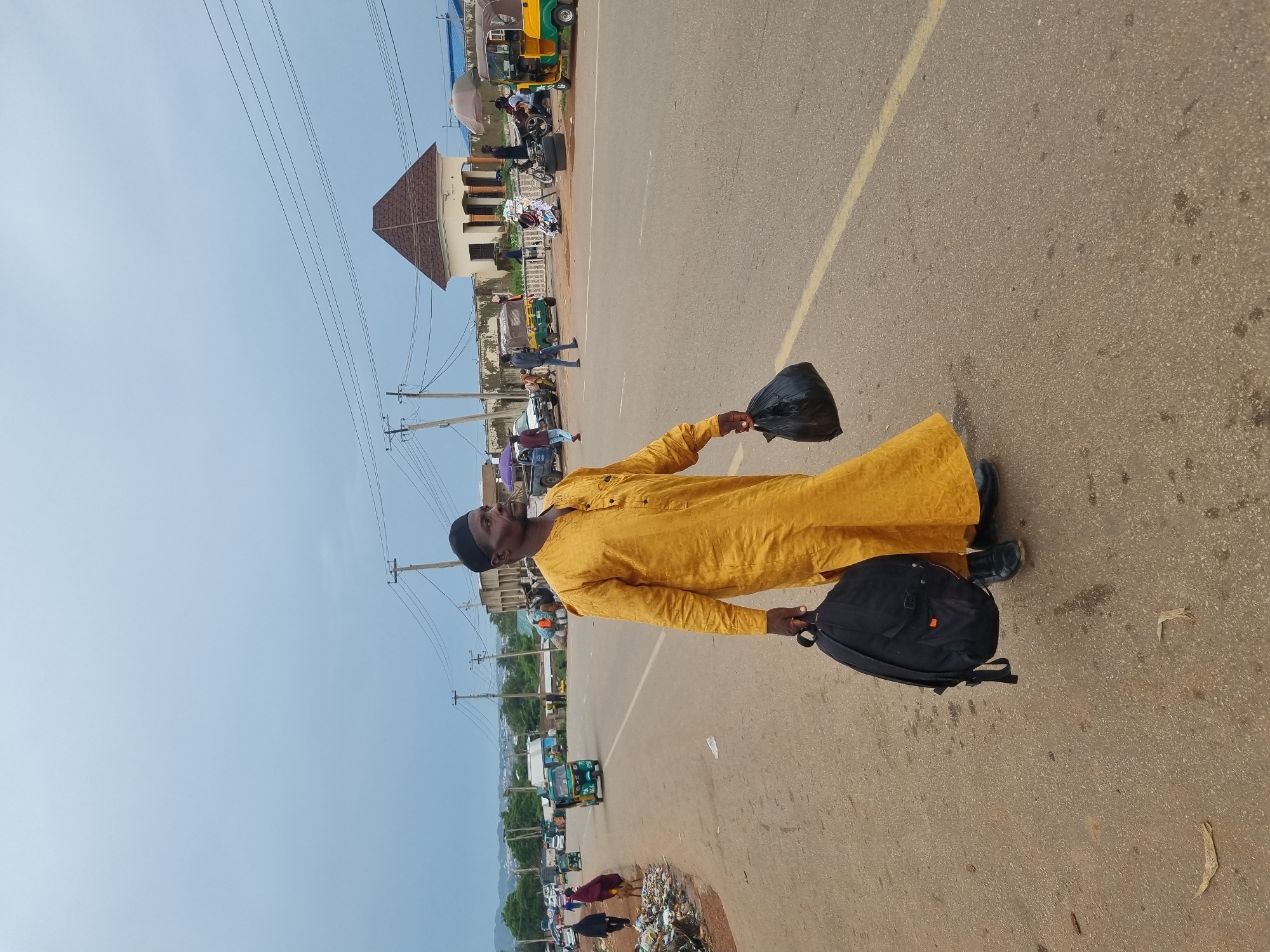 A man wearing a yellow traditional outfit and a black cap walks across a paved road in Jos, Nigeria. He is carrying a black backpack in his left hand and a black plastic bag in his right hand. The road is lined with a gutter on the left, filled with trash. In the background, there are several vehicles, including a green and yellow trishaw, a white pickup truck, and other cars. Buildings and utility poles are also visible. The sky is overcast.