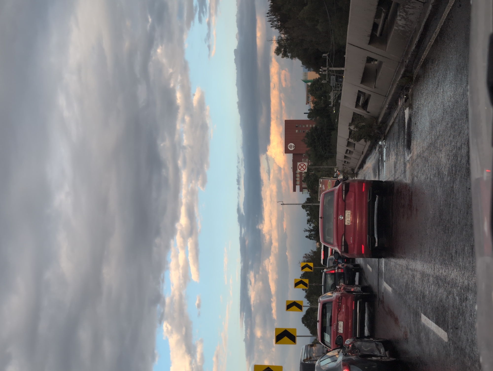 The image depicts a traffic jam on a wet road in Mexico City, Mexico. Multiple cars are visible, with a red Volkswagen Jetta being prominent in the foreground. The road is lined with yellow directional signs. On the left, a concrete barrier separates the road from trees. The sky is cloudy with a mix of gray and blue hues. A building with a “no U-turn” sign is visible in the distance, along with other buildings and trees. The image is oriented vertically.