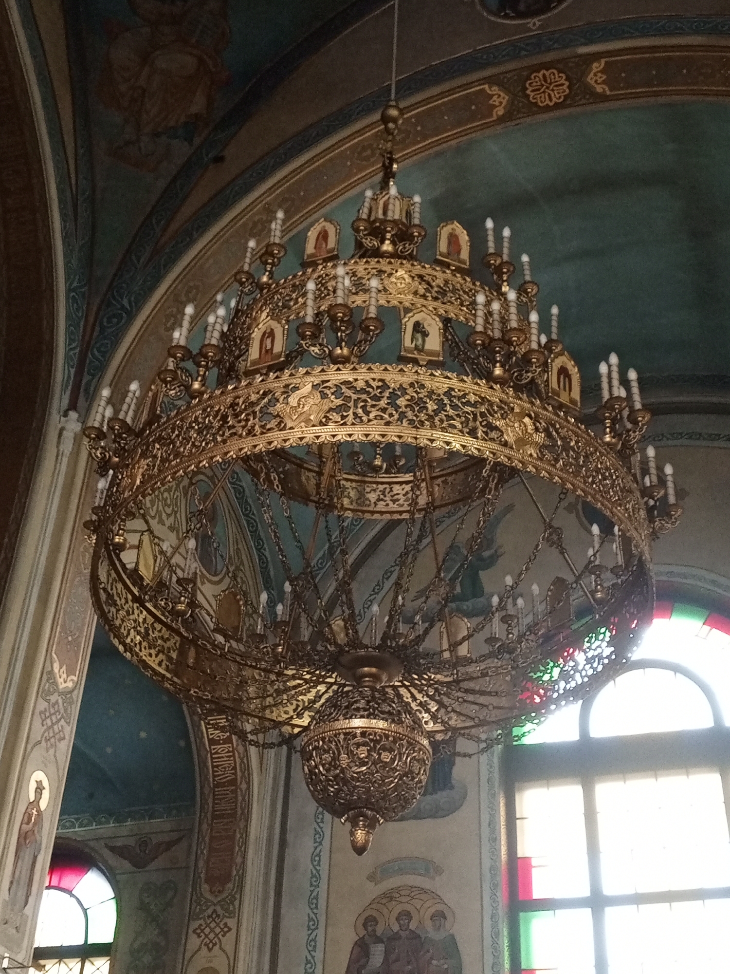 A grand, ornate golden chandelier hangs from the ceiling of a dimly lit church. The chandelier is multi-tiered and intricately detailed, with numerous candle-like light fixtures. The church interior features vaulted ceilings adorned with religious artwork, including frescoes and mosaics depicting figures in flowing robes. Arched windows with stained glass in red, green, and white are visible in the background. The overall atmosphere suggests a place of worship with rich historical and artistic significance.