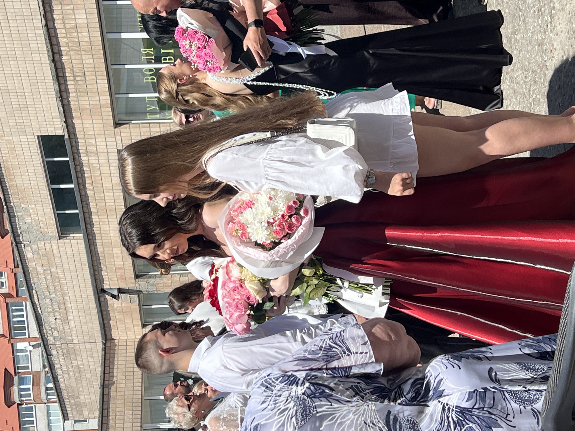 The image depicts a group of people, predominantly young women, at what appears to be a graduation ceremony or formal event, given the attire and the presence of bouquets. The setting is outdoors, with a brick building serving as a backdrop. The building has several windows and appears to be an institutional or residential structure. Sunlight suggests it is daytime, possibly late morning or early afternoon, casting shadows on the ground.
Several individuals are visible, mostly in the foreground and midground. One woman in a white dress is holding a bouquet of pink and white flowers. Another woman, wearing a long, dark dress, also has a pink bouquet. A third woman in a white dress with a quilted white purse is positioned further back. The image also shows the heads and shoulders of other people, some in formal wear, including a man in a white shirt. The atmosphere seems celebratory, with a focus on these individuals and their significant occasion.
One of the crops reveals text on the building, reading "ТУТ ВОЛЯ" which translates to "HERE IS FREEDOM" in Ukrainian. This text, along with the location context of Ternopil, Ukraine, strongly suggests the event is taking place in Ukraine. The presence of flowers, formal clothing, and the gathering of people indicates a special event, likely a graduation or celebration of achievement.