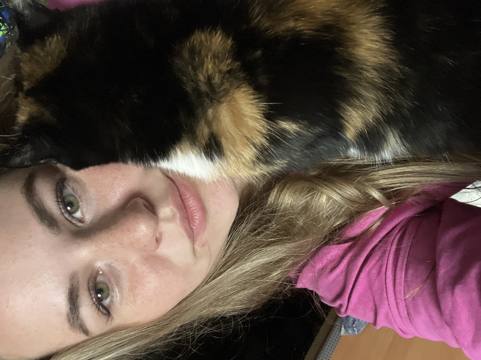 A woman with fair skin, green eyes, and blonde hair lies on her back. A calico cat with black, brown, and white fur rests on her face. The woman wears a pink shirt. The location is unknown and there is no evidence to support the assertion that this photo was taken in Chaikovsky, Russia.