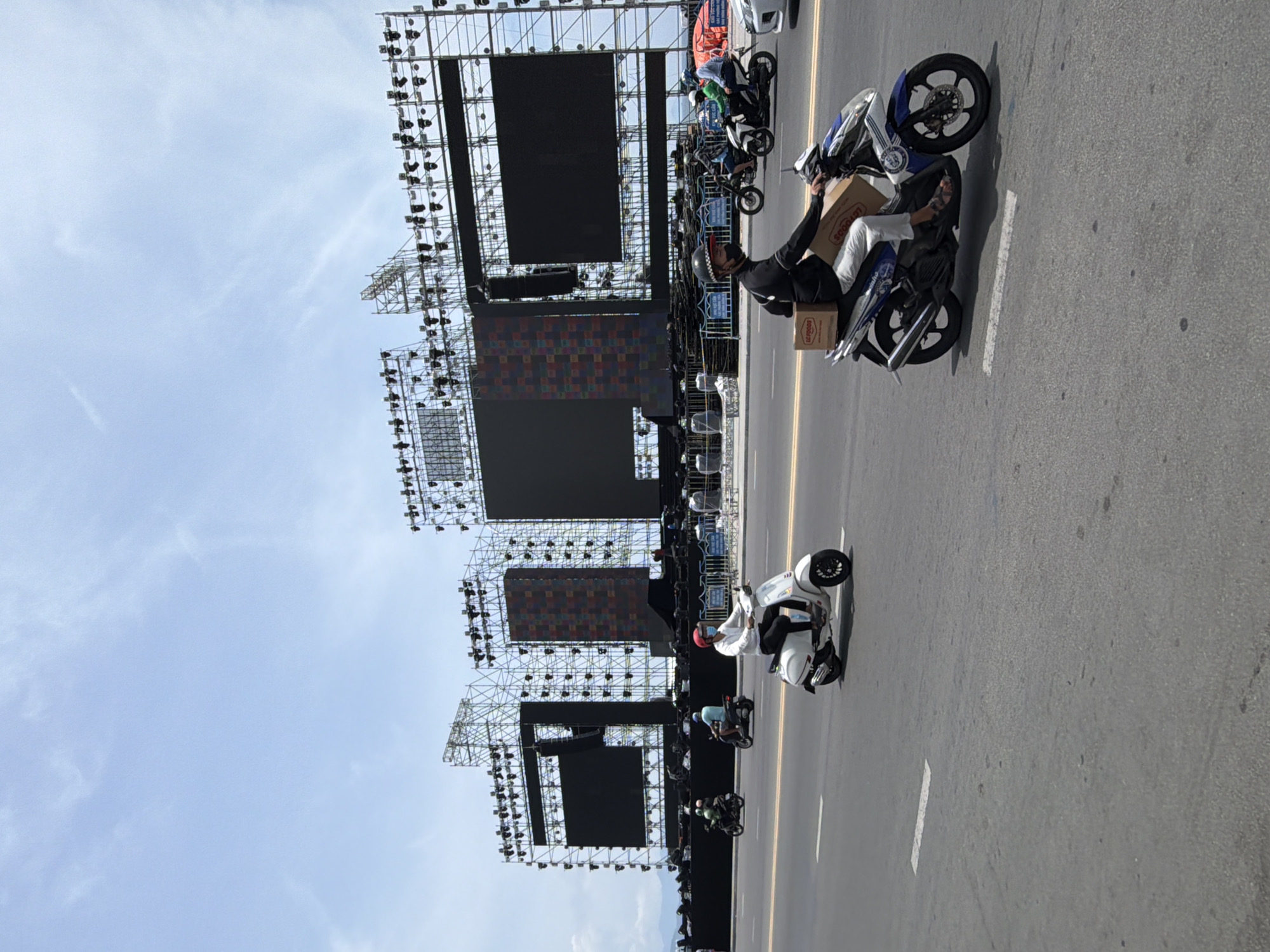 The image captures a street scene in Nha Trang Ward, Vietnam, with a large, multi-tiered stage structure dominating the background. The stage is elaborate, featuring extensive scaffolding and multiple large, dark screens. Several people are visible on the road, primarily riding motorbikes. In the foreground, a person on a white scooter, wearing a white shirt, black pants, and a red helmet with a face mask, is riding towards the viewer. Further back, another person on a similar white scooter is seen from the side, also wearing a helmet. To the right, a person on a blue and white motorbike, wearing a black jacket, helmet, and mask, is carrying two cardboard boxes on the back. The road is asphalt, with white lane markings. The sky is blue with scattered clouds. The overall impression is one of a busy urban environment with preparations for a large event.