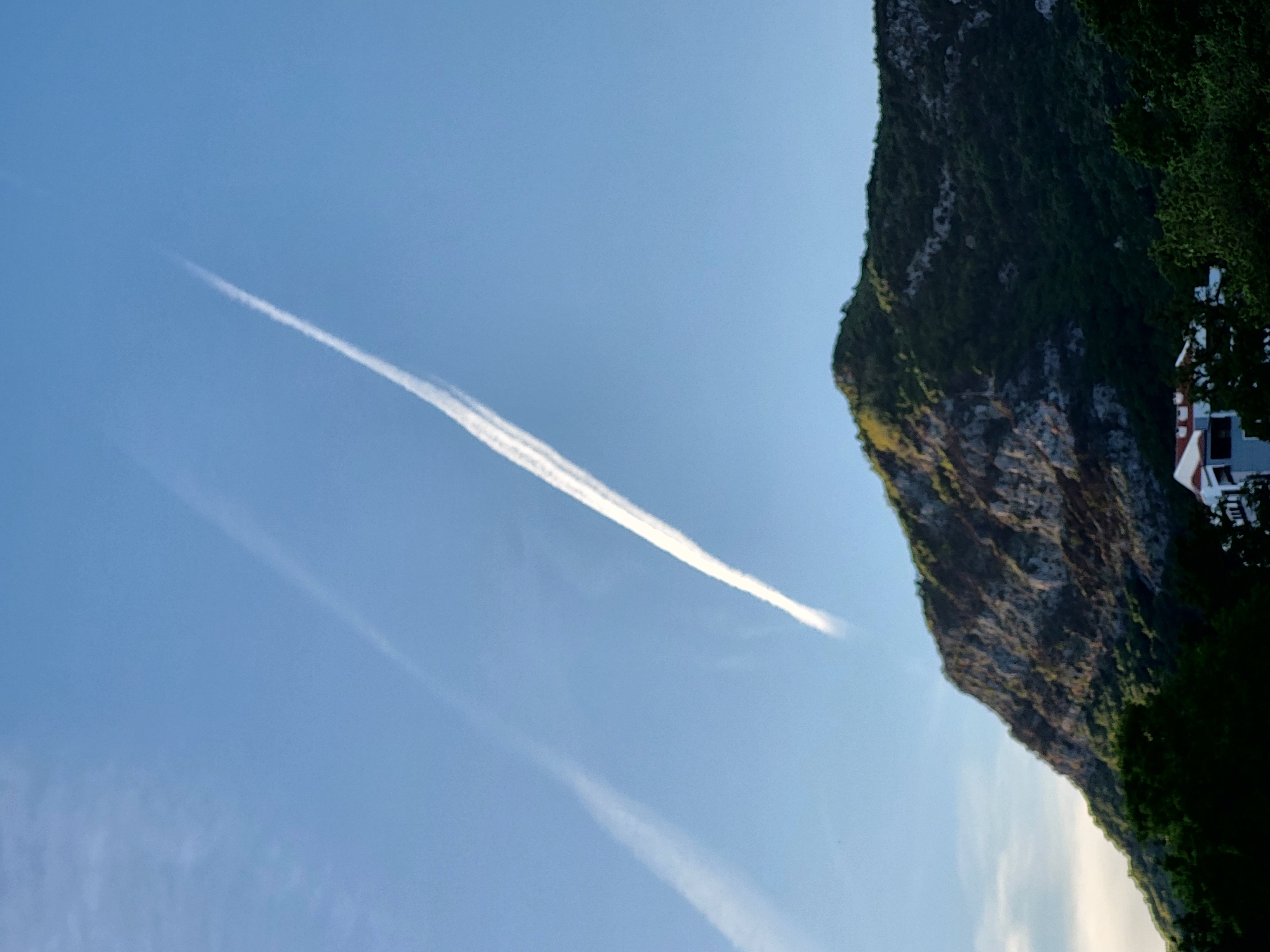 The image captures a clear blue sky dominating the left and upper portions of the frame, with a large mountain or cliff face on the right. The sky is a vivid, bright blue, indicating clear weather. Two white contrails are visible; a prominent, linear contrail stretches diagonally from the mid-left towards the upper right, and a fainter, wispy contrail appears below it, fading towards the center.
The right side of the image is occupied by a steep, rugged mountain or cliff covered in dense green vegetation. Rocky outcrops are visible through the foliage, showing variations in texture and color from grey to light brown, suggesting strong sunlight. At the very bottom right, partially obscured by trees, a small section of a white building with a reddish-brown roof is visible.
The scene depicts a sunny daytime, likely morning or early afternoon, given the bright illumination and cloudless sky. There is no explicit activity occurring, and no people are visible. The image conveys a sense of natural grandeur and open space. No text is visible in the frame. The landscape aligns with the characteristics of coastal and mountainous regions, specifically consistent with the provided location context of Budva, Montenegro.