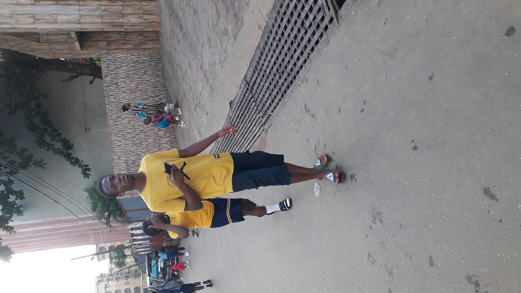The image shows a young man standing on a paved surface, likely a street or sidewalk, in Onitsha, Nigeria. He is wearing a yellow shirt and dark shorts, and he is holding a mobile phone in his left hand. Another person, also in a yellow shirt and dark shorts, is standing next to him, partially obscured.
A large metal drain grate is located adjacent to them. In the background, other people and vehicles are visible. Buildings and trees line the street, providing context for an urban environment. A specific Onitsha landmark is not identifiable from the image.