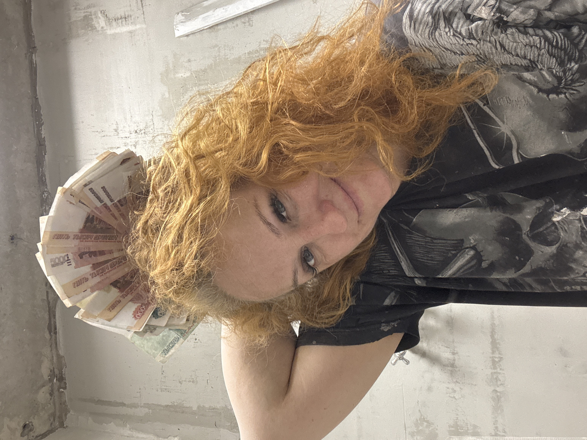 The image, oriented sideways, presents a close-up portrait of a woman with vibrant red, curly hair, looking directly at the camera with a subtle smile. Her light-colored eyes are prominent, and she appears to be wearing a dark-colored t-shirt with abstract grey and white patterns. Her right arm is bent, with her elbow visible in the lower portion of the frame. In her left hand, fanned out near her hair, she displays a collection of Russian ruble banknotes. The visible currency includes several 5000 ruble notes, clearly showing the text "ПЯТЬ ТЫСЯЧ РУБЛЕЙ" (Five Thousand Rubles), and at least one 1000 ruble note with "ТЫСЯЧА РУБЛЕЙ" (One Thousand Rubles) visible. The background consists of an unfinished or roughly plastered concrete wall, indicating an indoor setting, possibly a room undergoing renovation or construction in Chaikovsky, Russia. The lighting appears bright and uniform, typical of indoor illumination. The woman's expression suggests confidence or satisfaction while presenting the money.