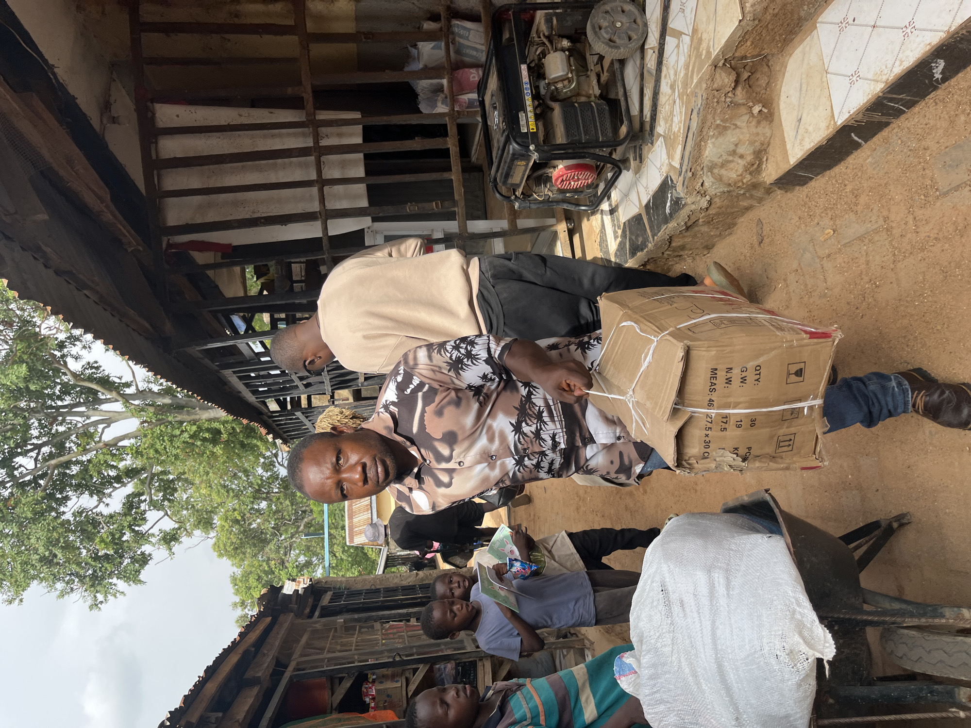 In an outdoor marketplace in Jos, Nigeria, a man in a patterned shirt carries a cardboard box. He is surrounded by other people, including children holding books. To the right, a generator is visible. The ground is dirt, and structures of corrugated metal and wood line the market. The scene appears to be during daylight.