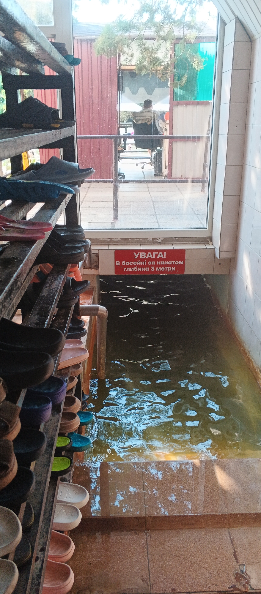 The image depicts a swimming pool area in Berehove, Ukraine, as indicated by the red sign with Cyrillic text that reads "УВАГА! В басейні за канатом глибина 3 метри," which translates to "ATTENTION! The depth of the pool behind the rope is 3 meters." The foreground features a multi-tiered rack filled with various types of footwear, primarily flip-flops and sandals, in an assortment of colors including black, blue, pink, and white. The rack appears to be made of metal and wood. Adjacent to the shoe rack is a section of a swimming pool filled with dark, rippling water. A metal pipe is visible entering the water. In the background, through a large window, a person is seated at a desk in an office-like setting, with some green foliage visible outside. The overall scene suggests an indoor or semi-outdoor swimming facility with changing room amenities.